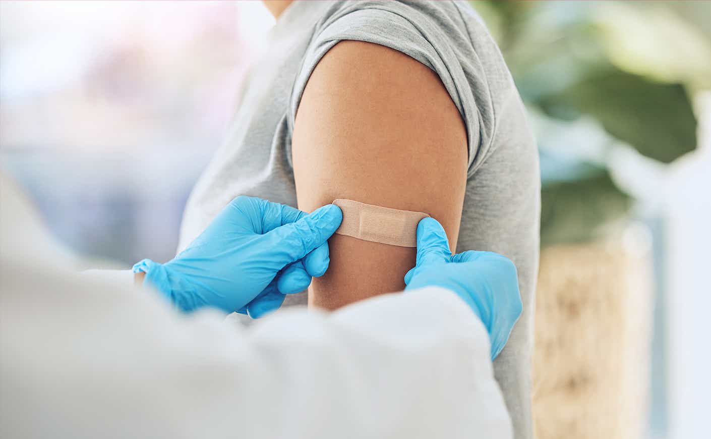 Gloved hands pressing bandaid onto person's arm, like after they got a vaccine.