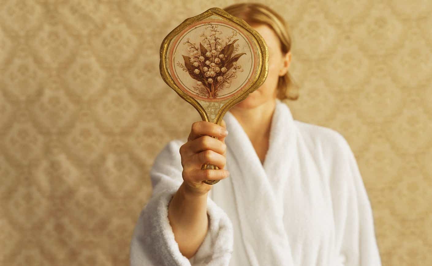 woman in a bathrobe looking at her reflection in a hand mirror