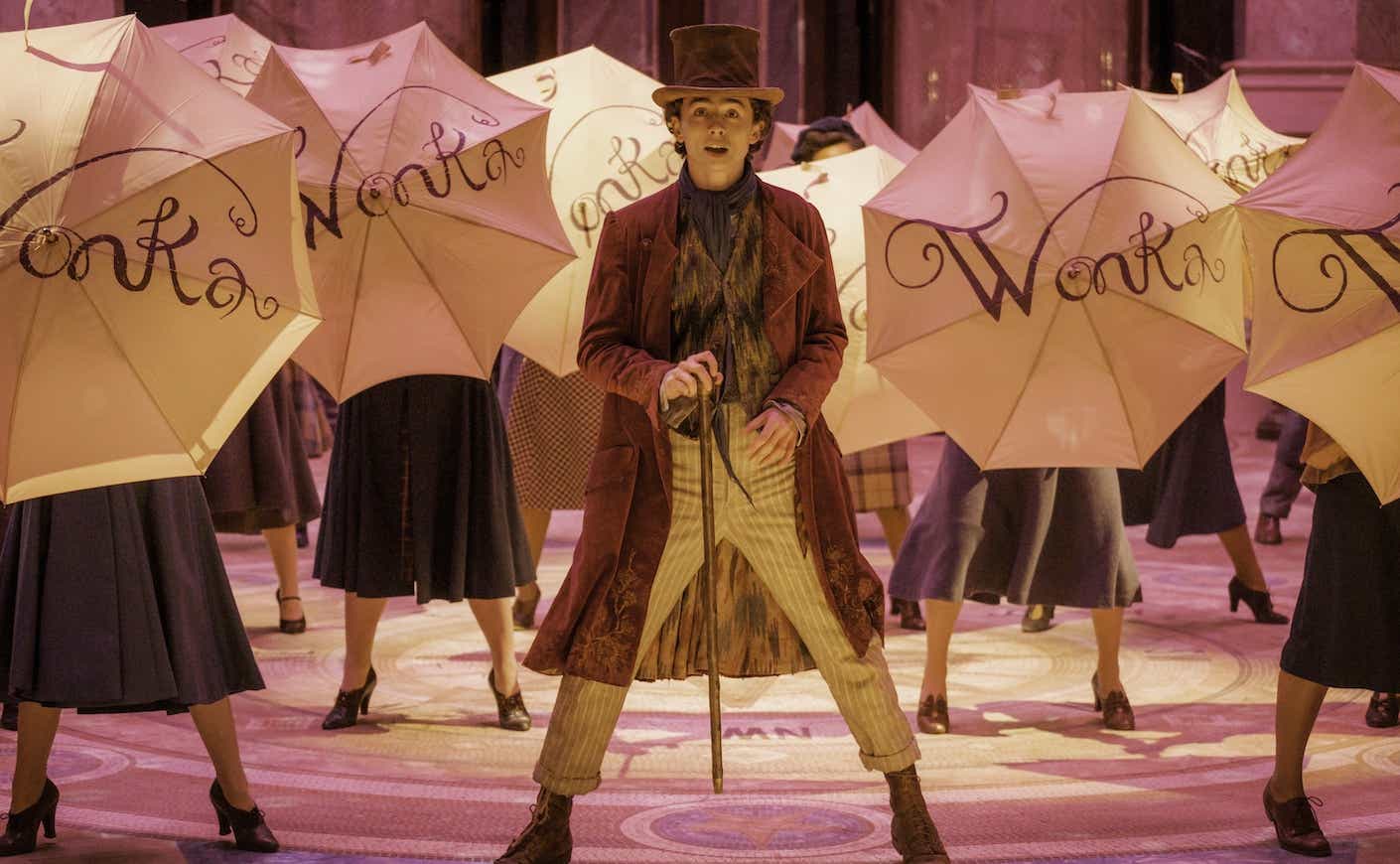timothee chalamet on stage as willy wonka surrounded by people holding umbrellas that say "wonka"