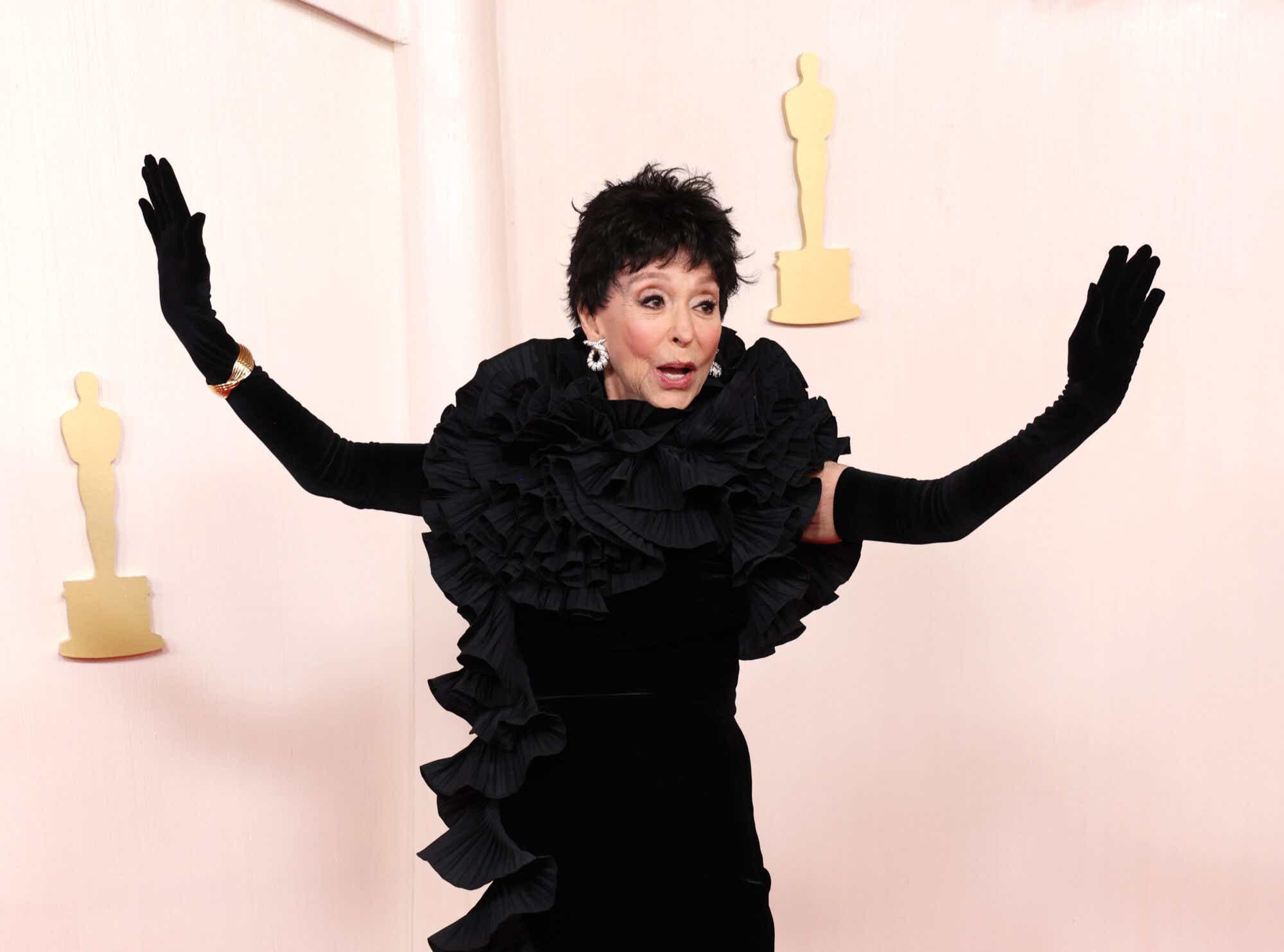 Rita Moreno attends the Oscars wearing a black town with ruffling around her shoulders and down the side and matching black gloves.