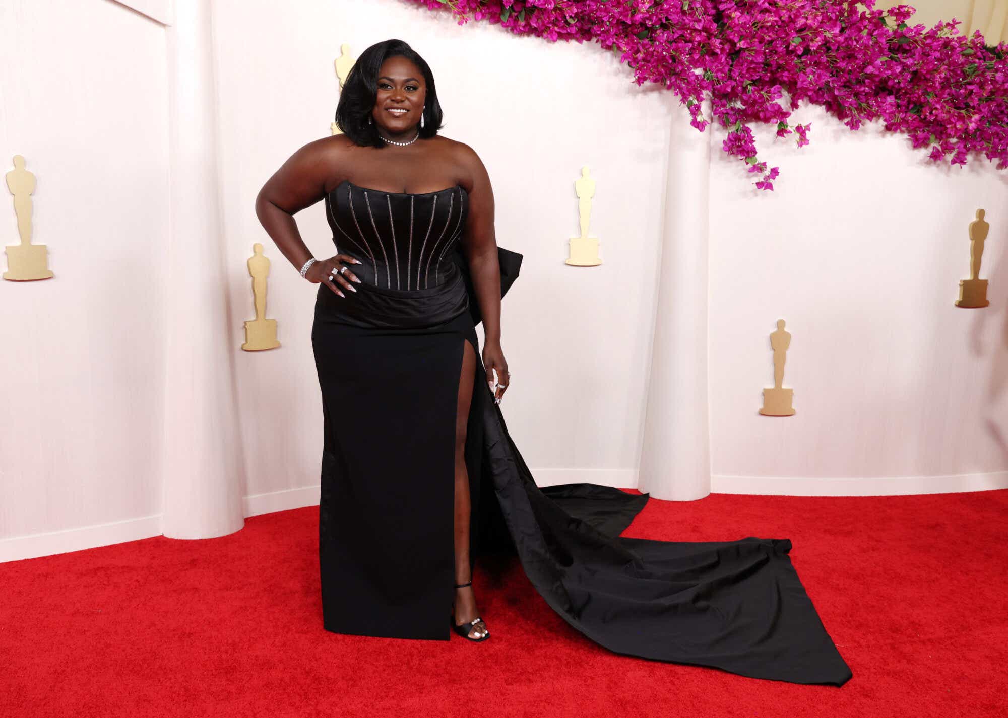 Danielle Brooks arrives at the Oscars wearing a black gown with a beaded corset top, daring leg slit, and a train.