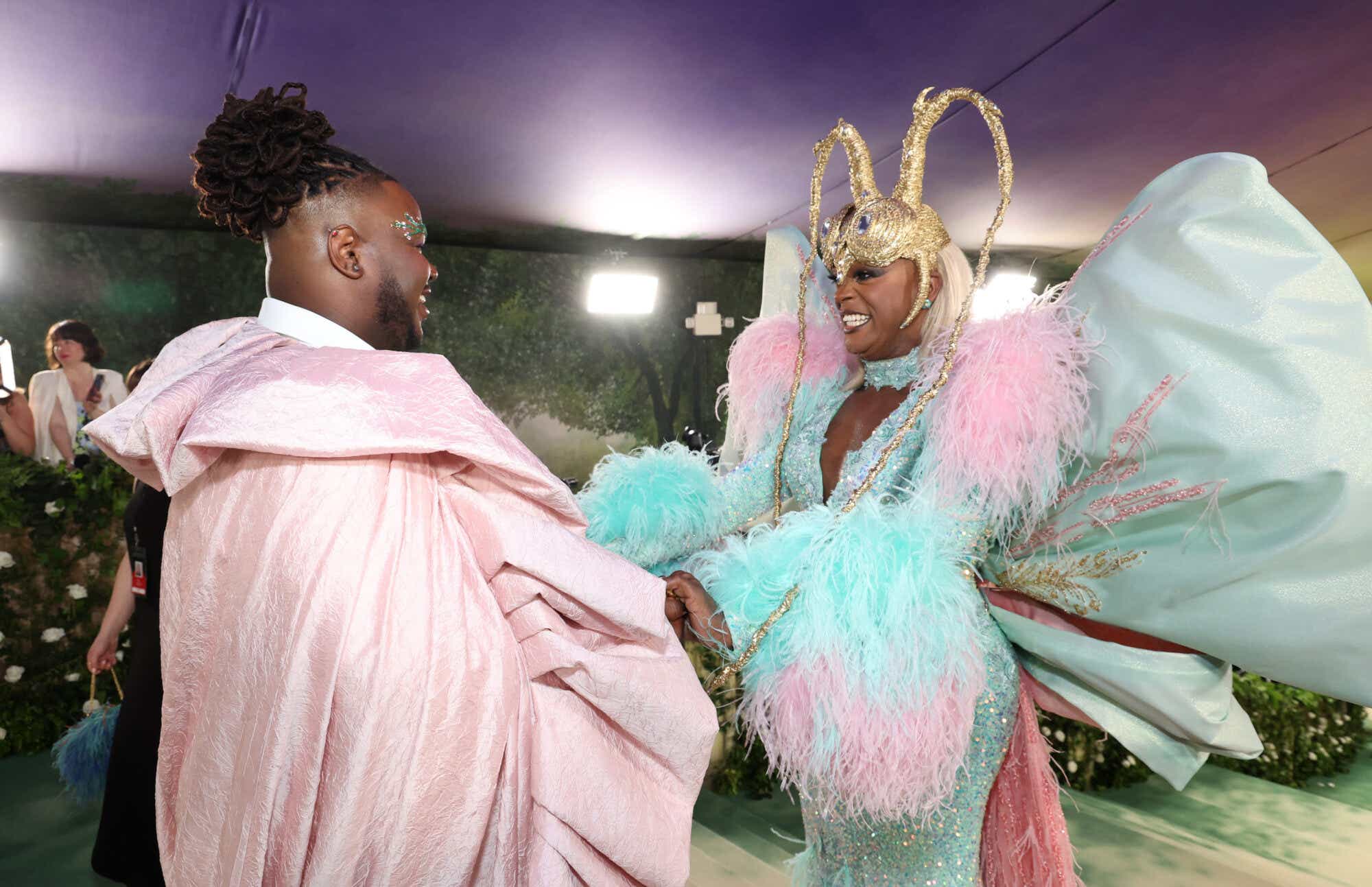 Kyle Ramar Freeman wears a pale pink cape with a large collar and bedazzled eyebrows, and J. Harrison Ghee wears a turquoise and pink dress with diamante detail and a Maleficent-style gold headdress