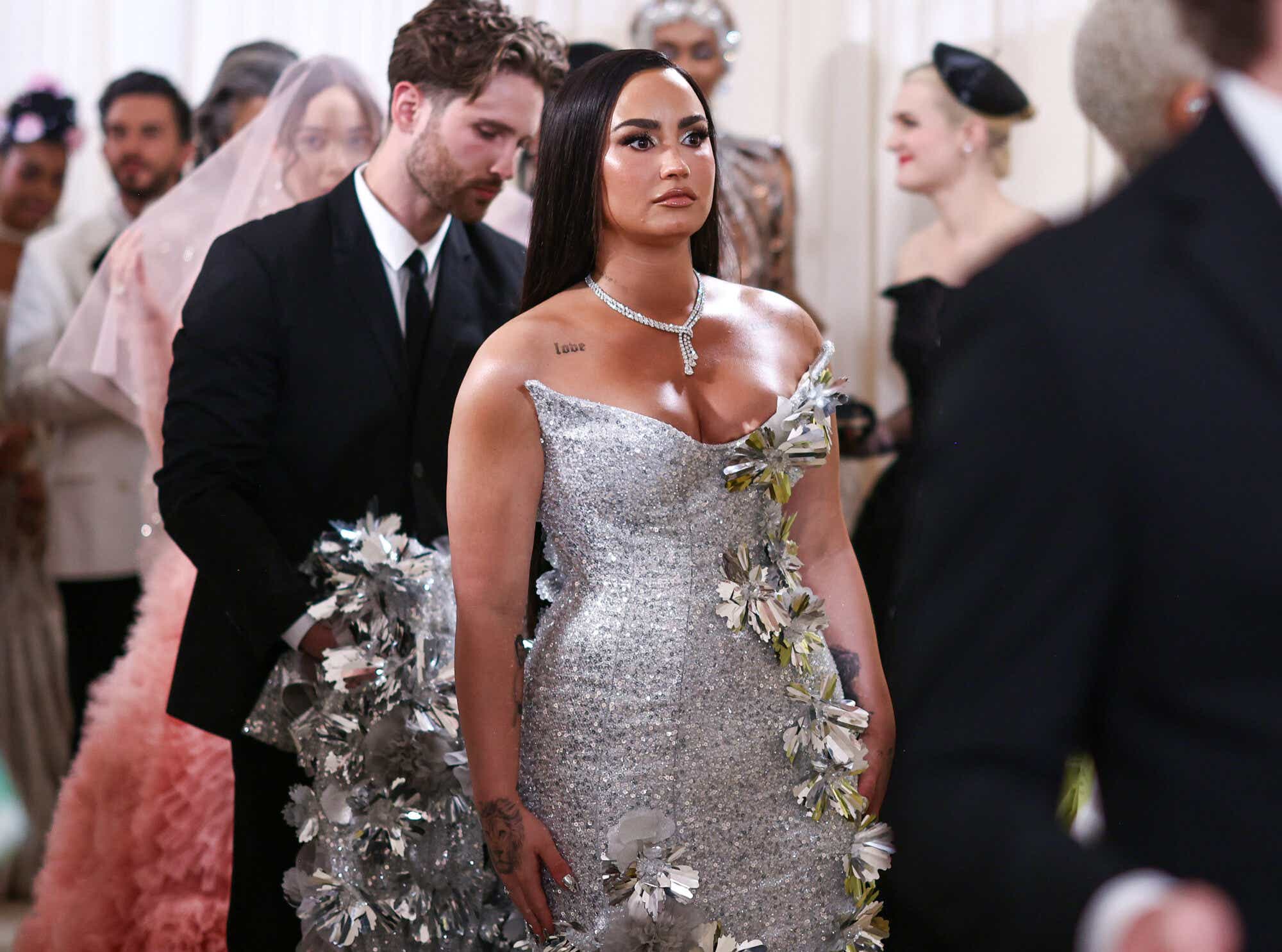 Demi Lovato wears a strapless silver dress with a structured neckline and delicate metallic flowers trailing down one side, merging into a flowery train