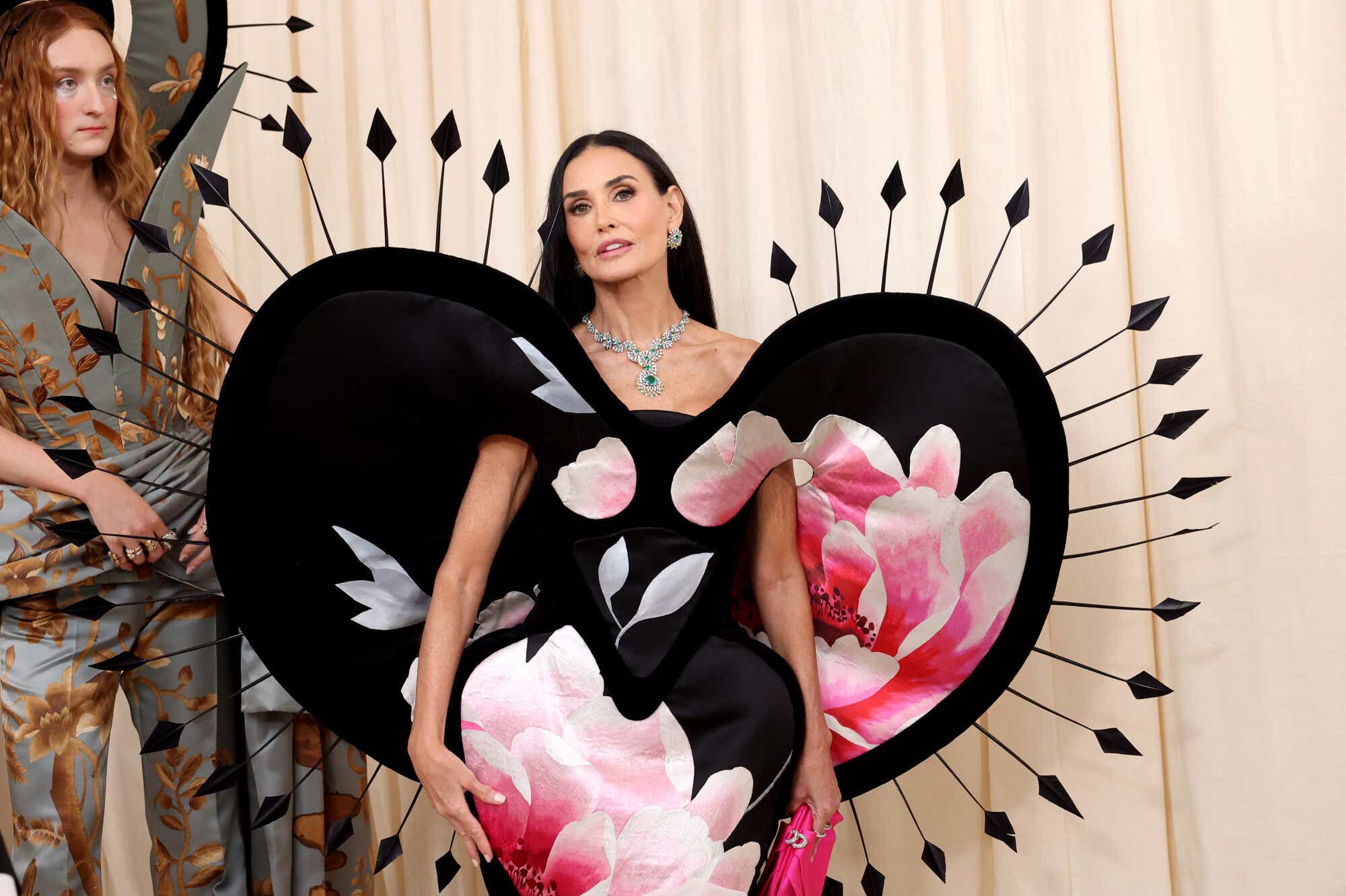 Demi wears a black dress with a pale pink rose design and an exaggerated structured hip, with a stiff heart-shaped cape trimmed with black arrows. 