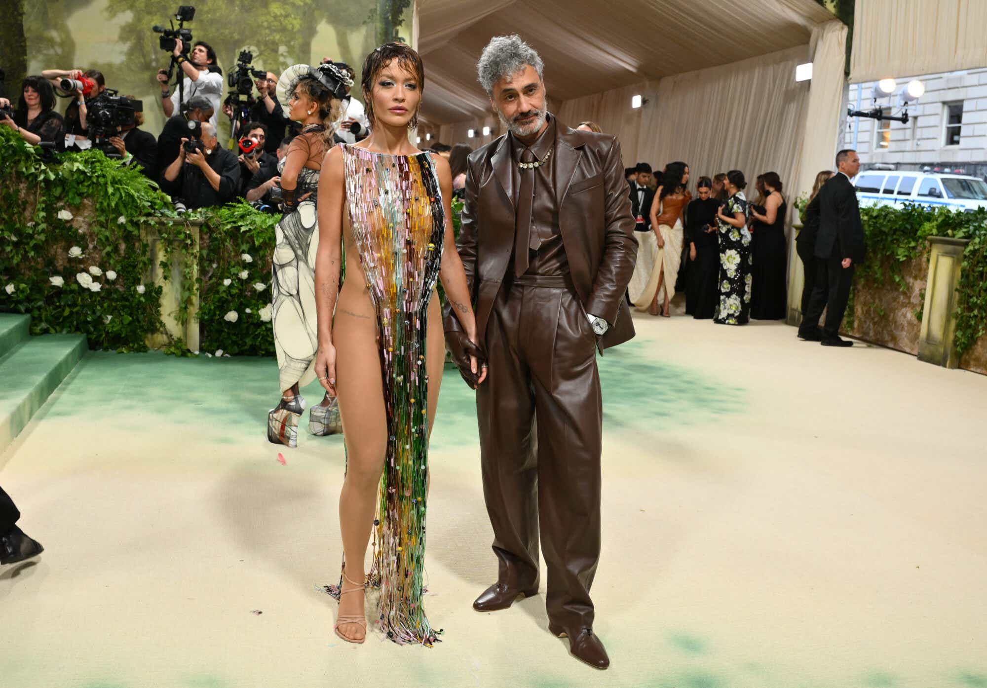 Rita Ora wears a multicolored beaded gown draped around her neck — and nothing else. Taika Waititi is in a brown leather suit jacket, pants, shirt, and tie.