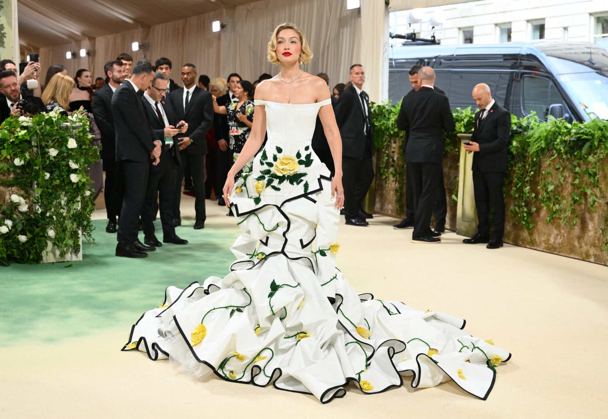 Gigi Hadid wears a bold red lip and her hair in pin curls, plus a white off-the-shoulder gown with yellow flowers and black trim.