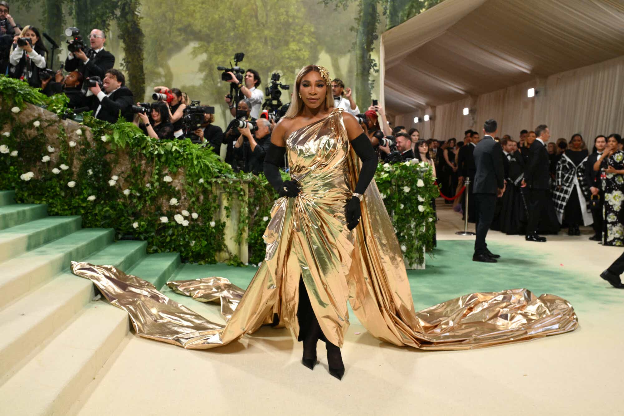 Serena Williams stuns in a shiny gold one-shoulder Grecian inspired gown with a cape and long train.