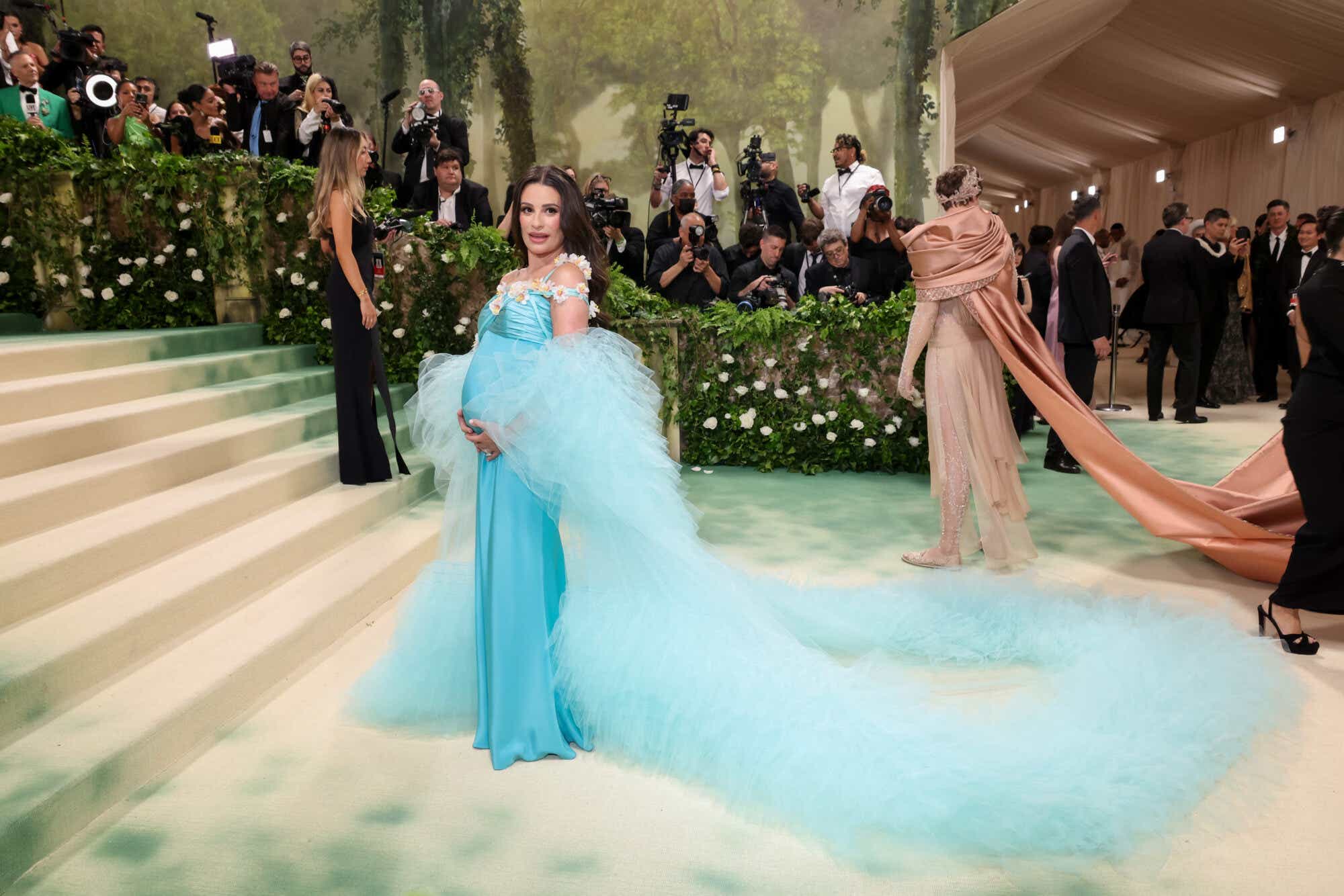 Lea Michele shows off her baby bump on the 2024 Met Gala red carpet, in a flowy turquoise dress with a dramatic train and daisys along the neckline.