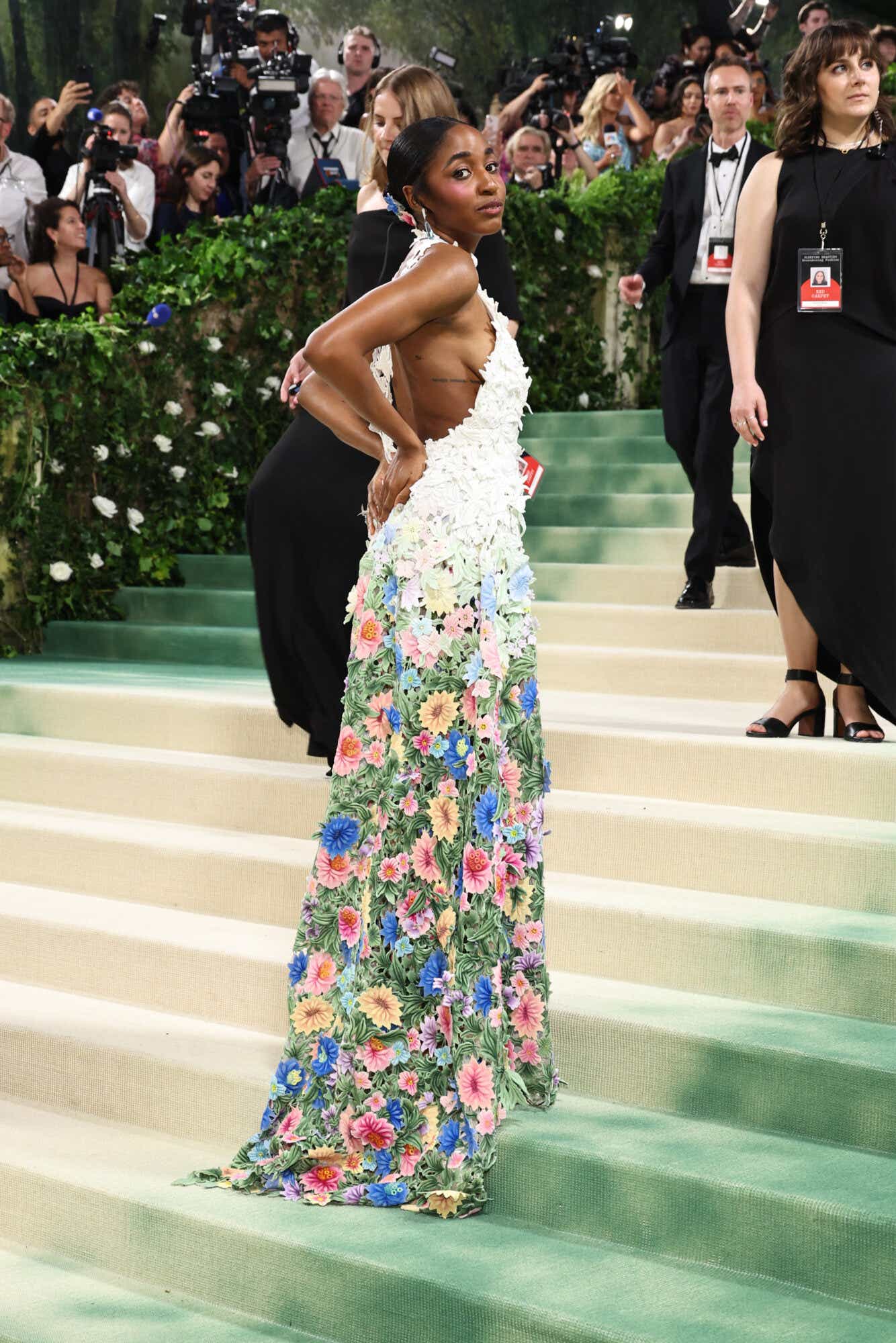 Ayo Edebiri wears a dress with floral appliques and a high neck. From the top it's white, towards the bottom the flowers are vibrant and multicolored.