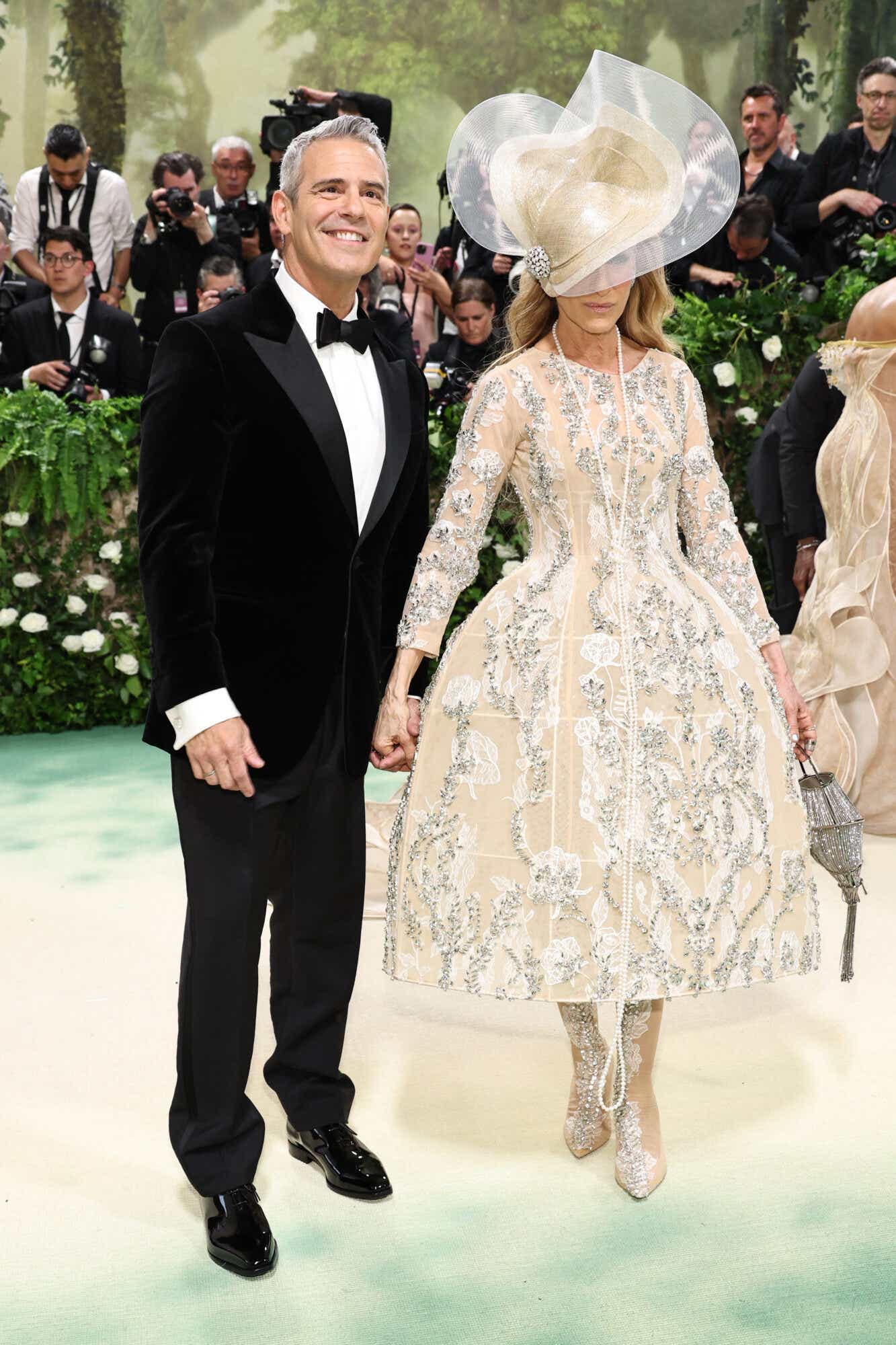 Andy Cohen wears a black tux. Sarah Jessica Parker wears an elaborately embroidered cream tea-length gown with a matching fascinator that covers one of her eyes.