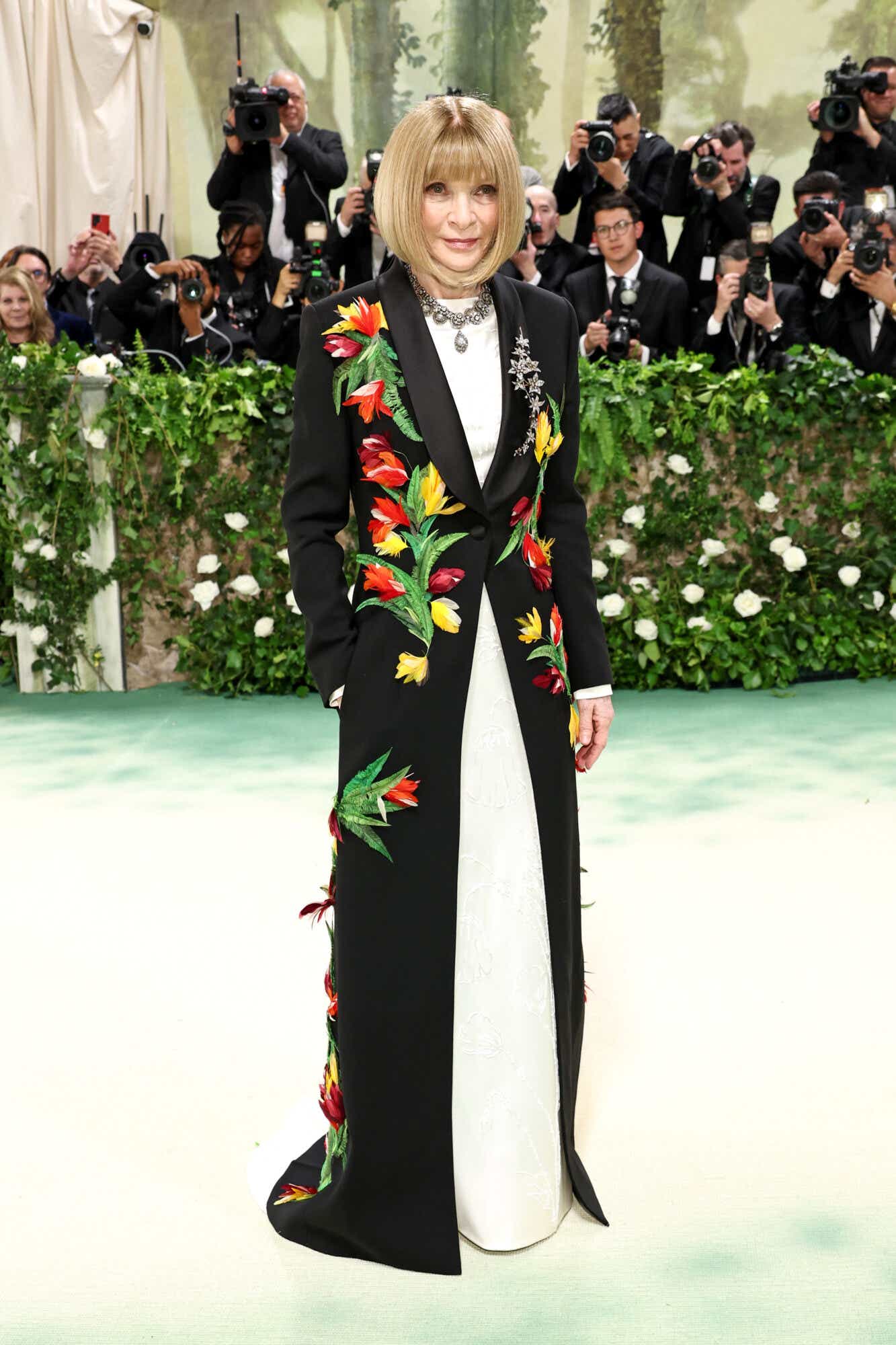Anna Wintour wears a white gown with a long black tuxedo jacket over it, adorned with red and yellow tulips.