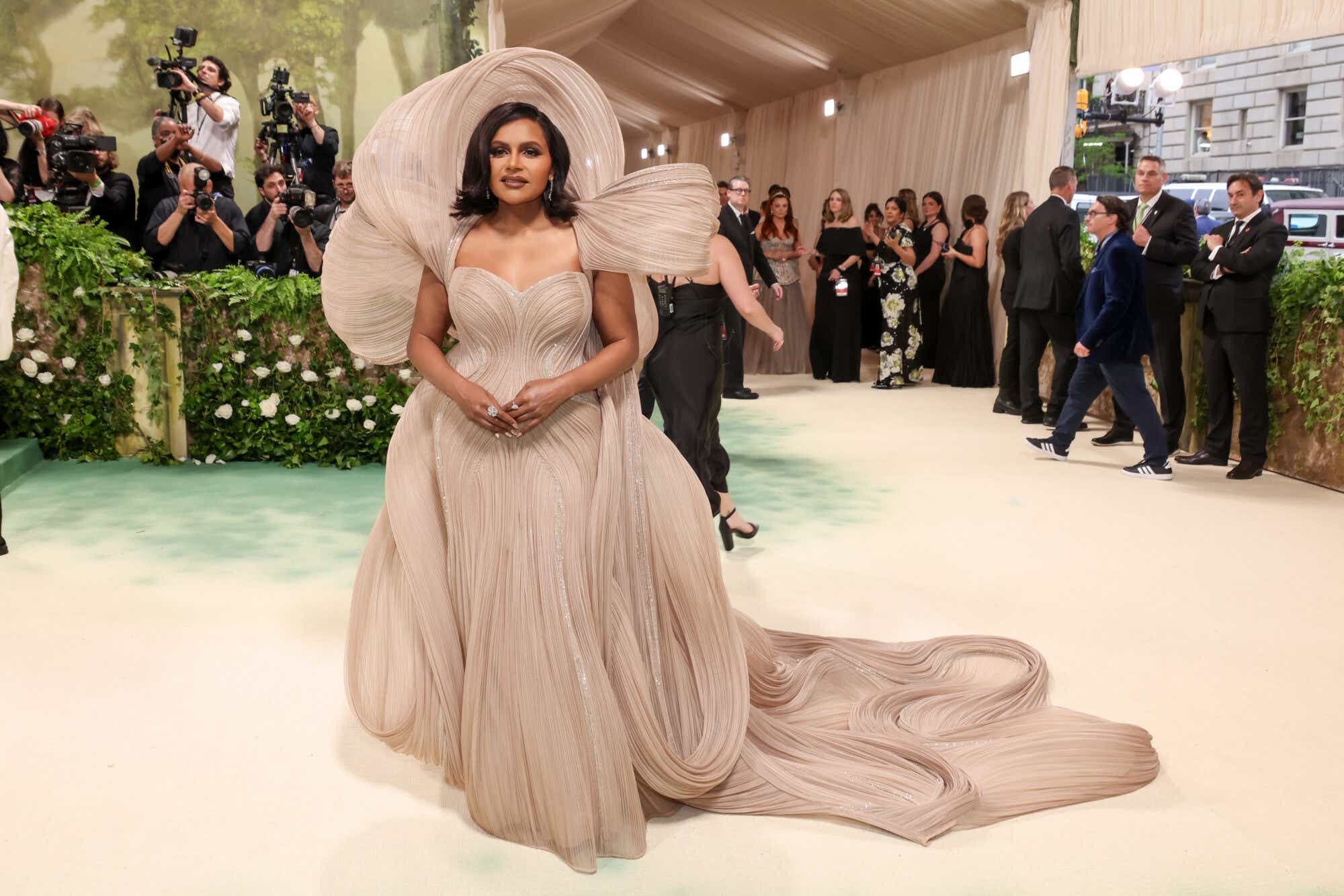 Mindy Kaling wears a beige dress with an ethereal over-the-head shoulder piece that matches the elegant draping of her gown.