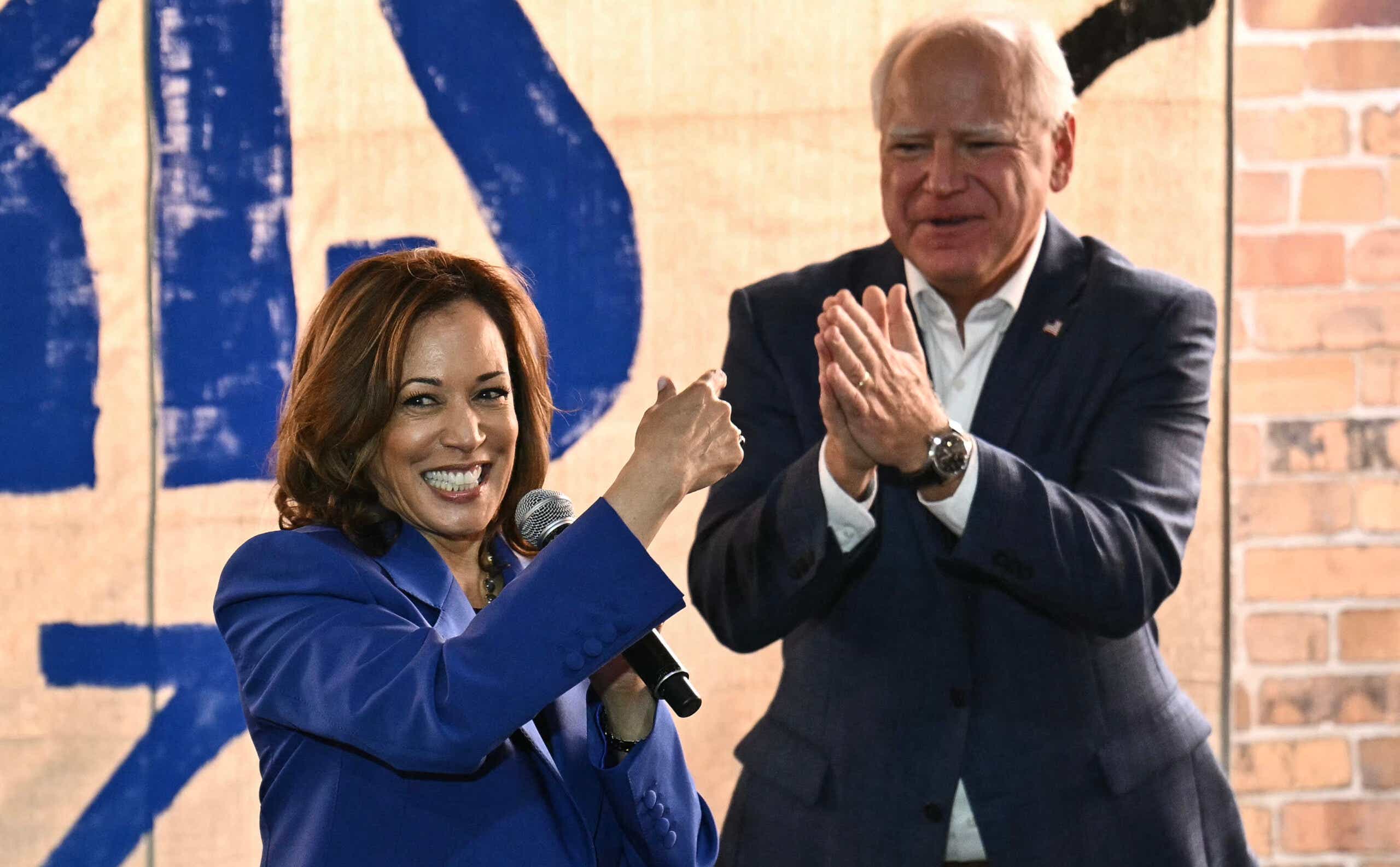 Kamala Harris holding a microphone, Tim Walz clapping behind her