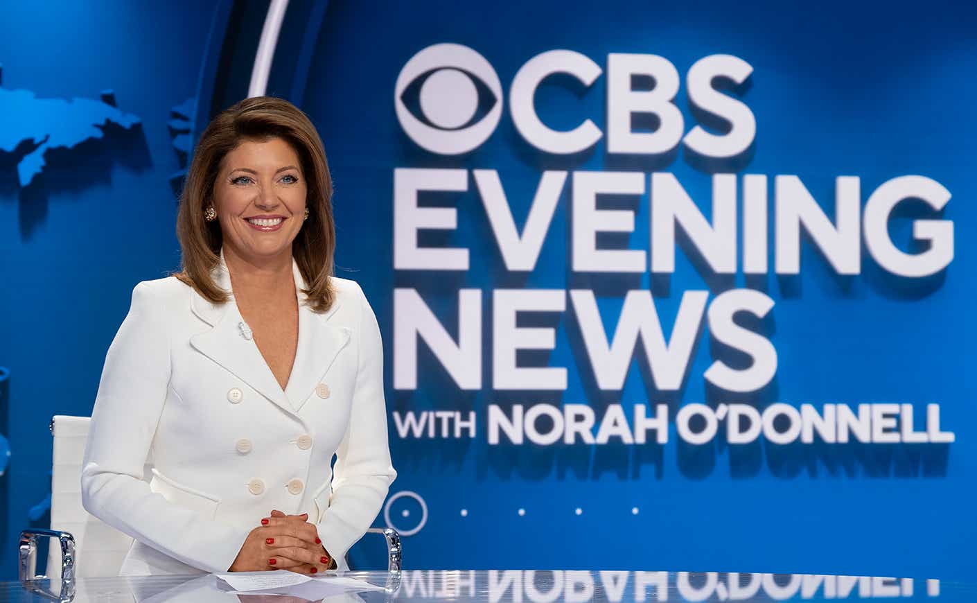 Norah O'Donnell in a white suit sitting at the anchor desk with the words "CBS Evening News with Norah O'Donnell" behind her