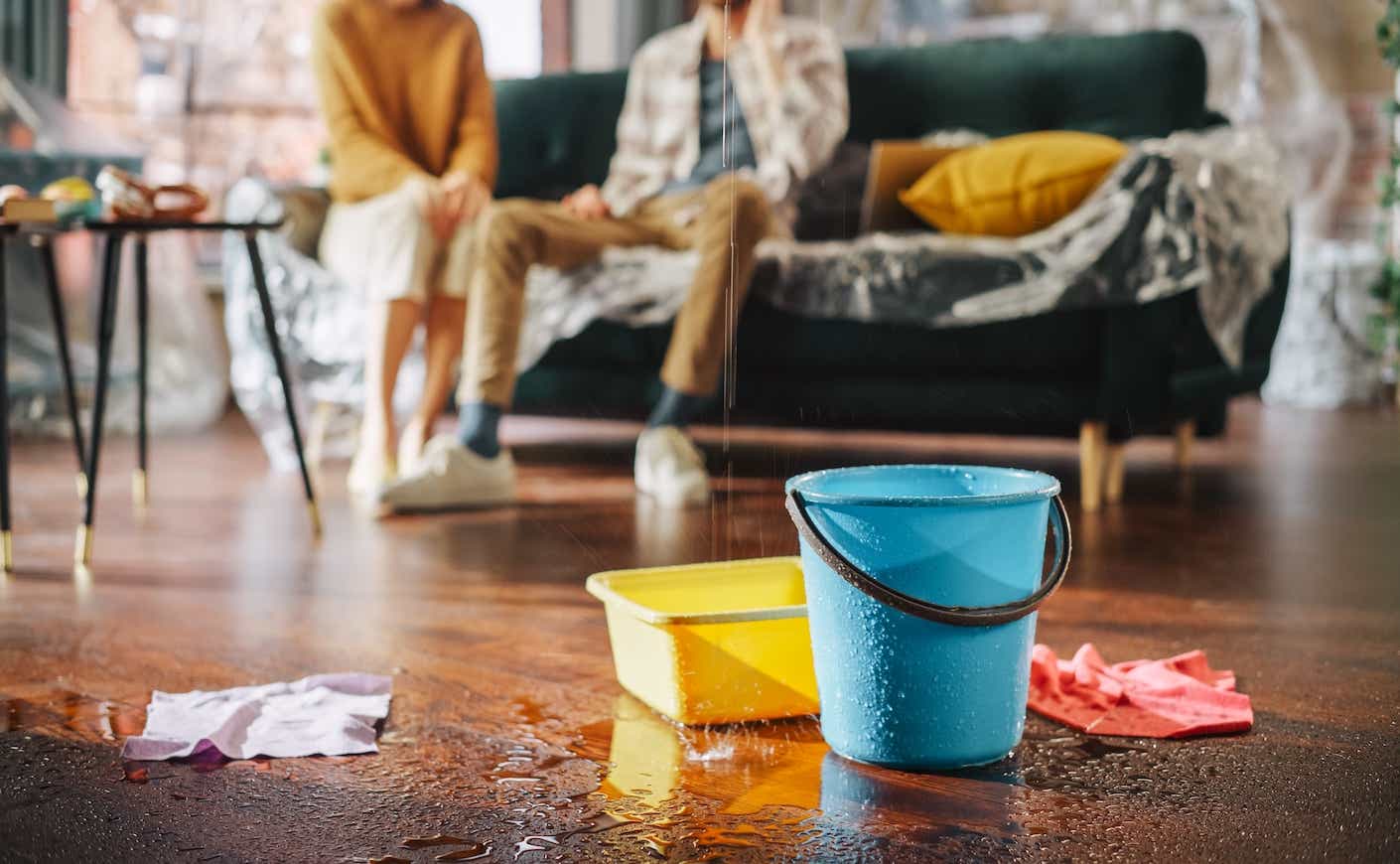 Water Drips into Buckets in Living Room. Angry Couple in Background Calling Insurance Company