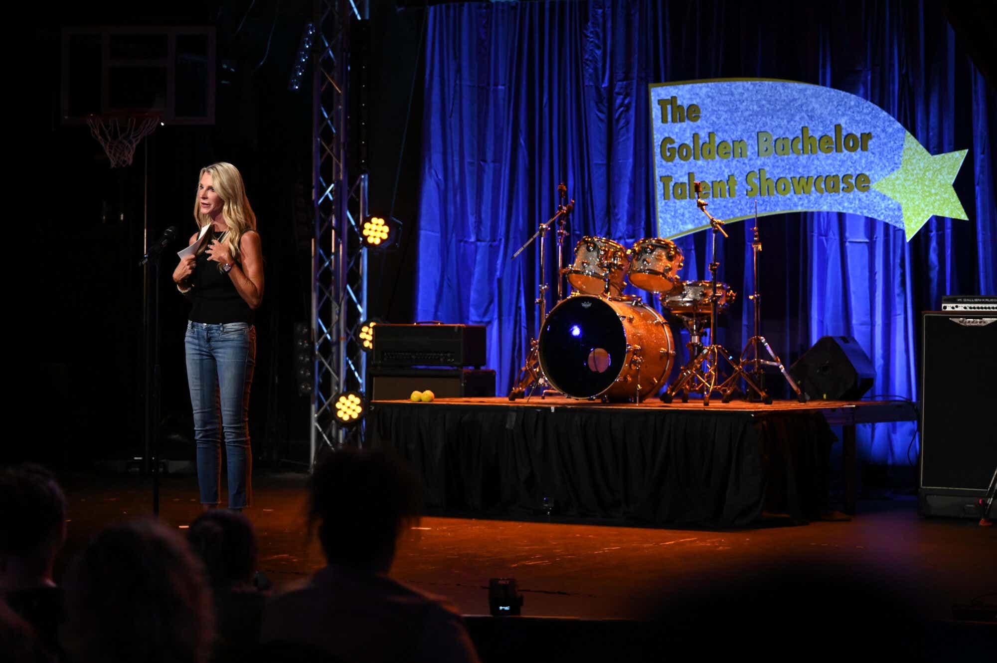 Joan Vassos in a black tank and jeans speaking into a microphone at the Golden Bachelor Talent Showcase