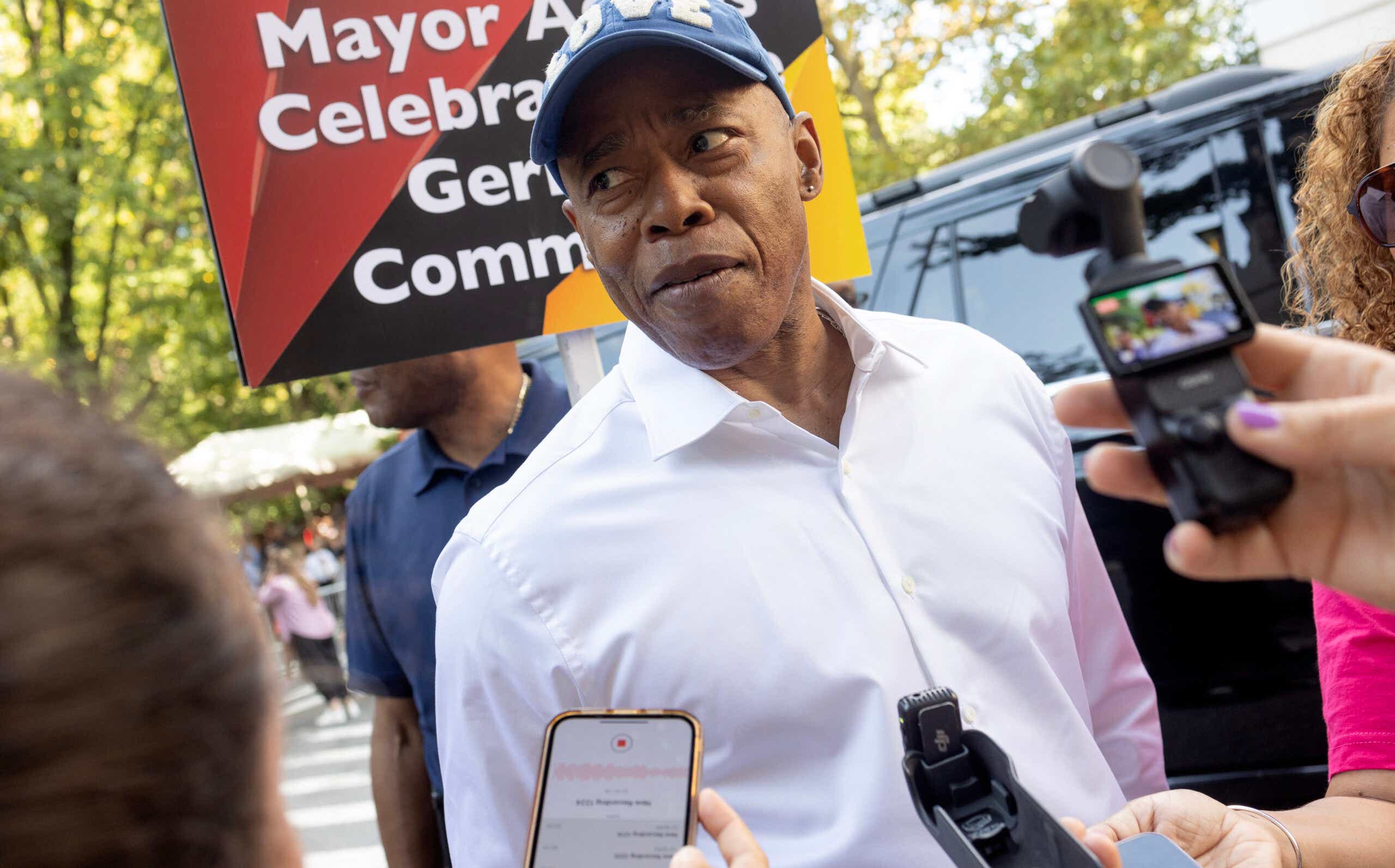 Eric Adams wearing a white shirt and a baseball cap being questioned by reporters