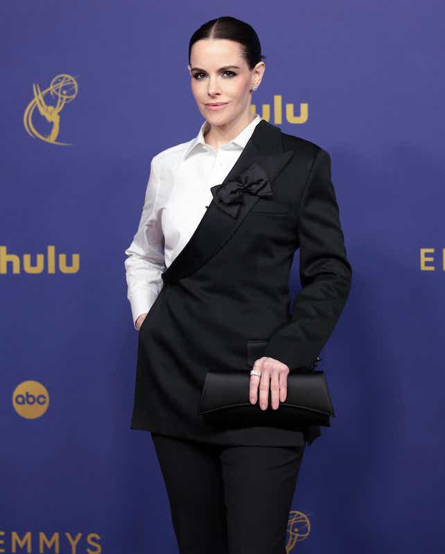 Emily Hampshire from Schitt's Creek is wearing a suit and pants. The jacket is colorblocked black and white across a diagonal, and there's a bowtie affixed to the lapel.