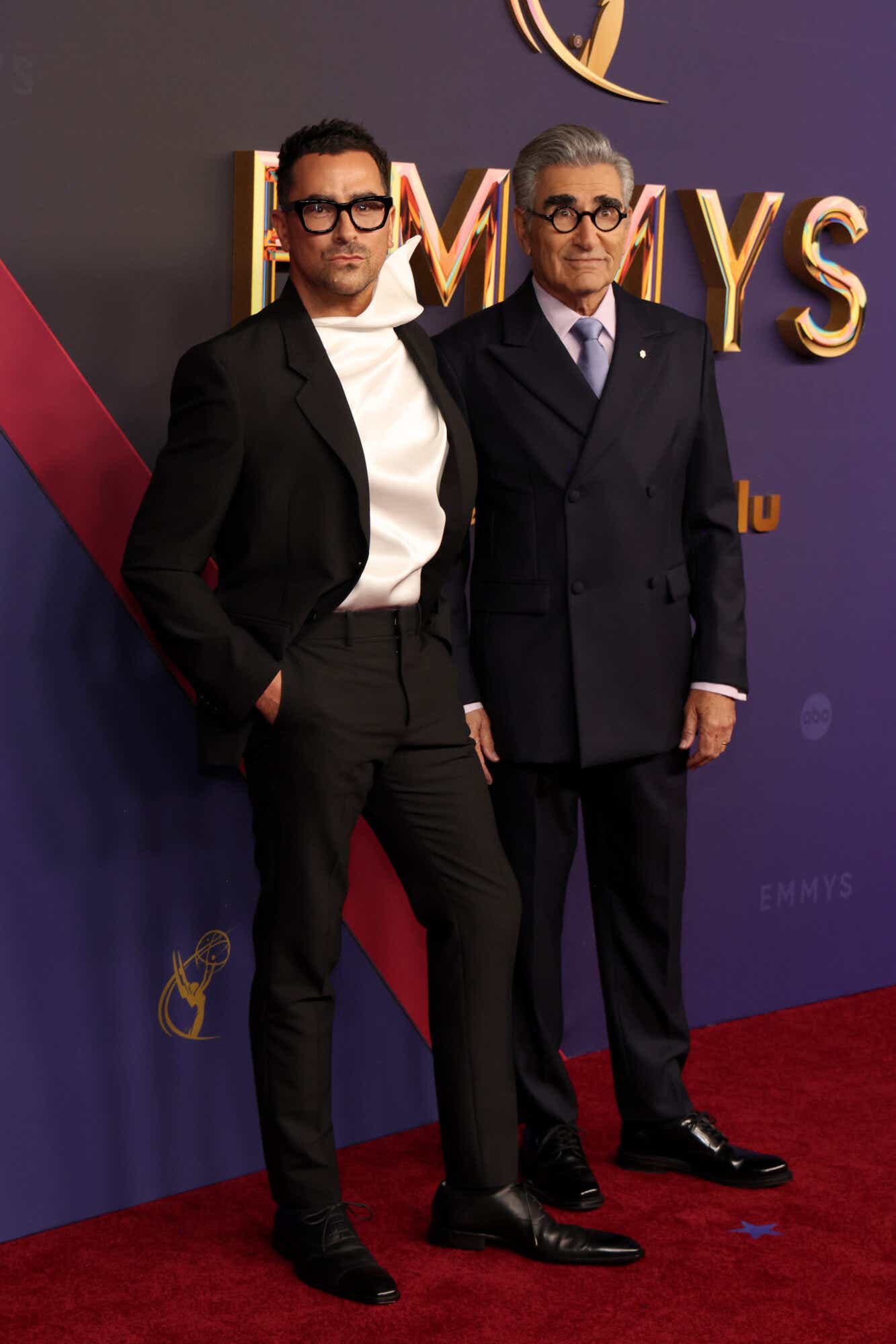 Dan Levy wears a black suit with a white top with a dramatic, architectural neck. Eugene Levy wears his signature round black-rimmed glasses and a navy suit with a light purple shirt.