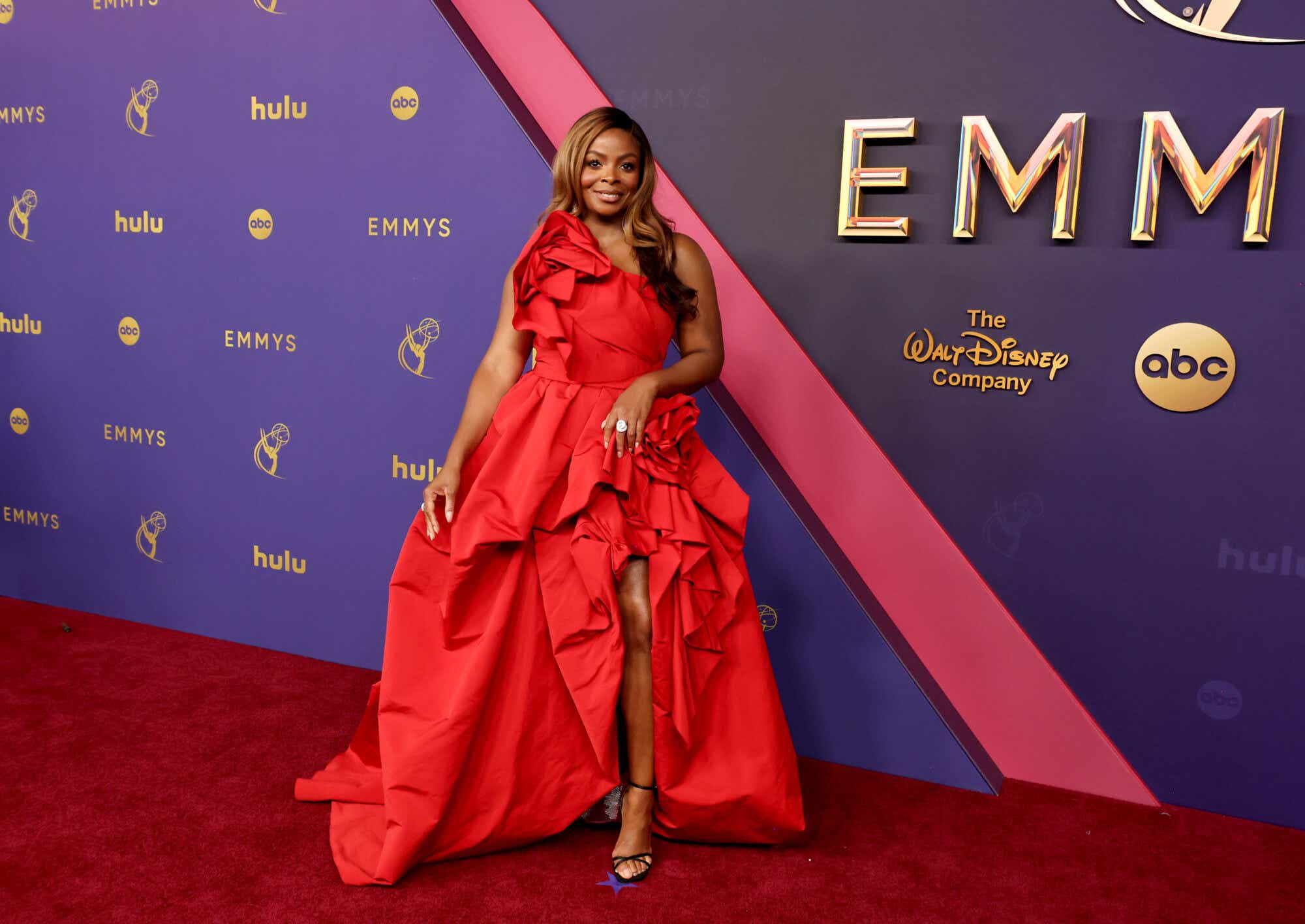 Janelle James wears a one-shoulder red gown with a dramatic, flowing skirt and rose details on the shoulder and hip.