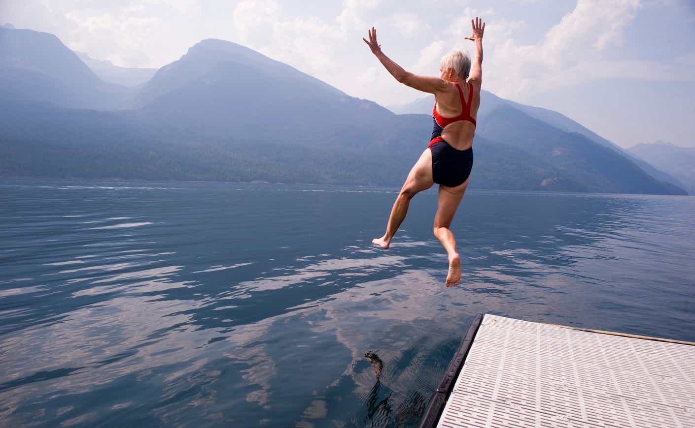 older woman jumping into a body of water surrounded by mountains