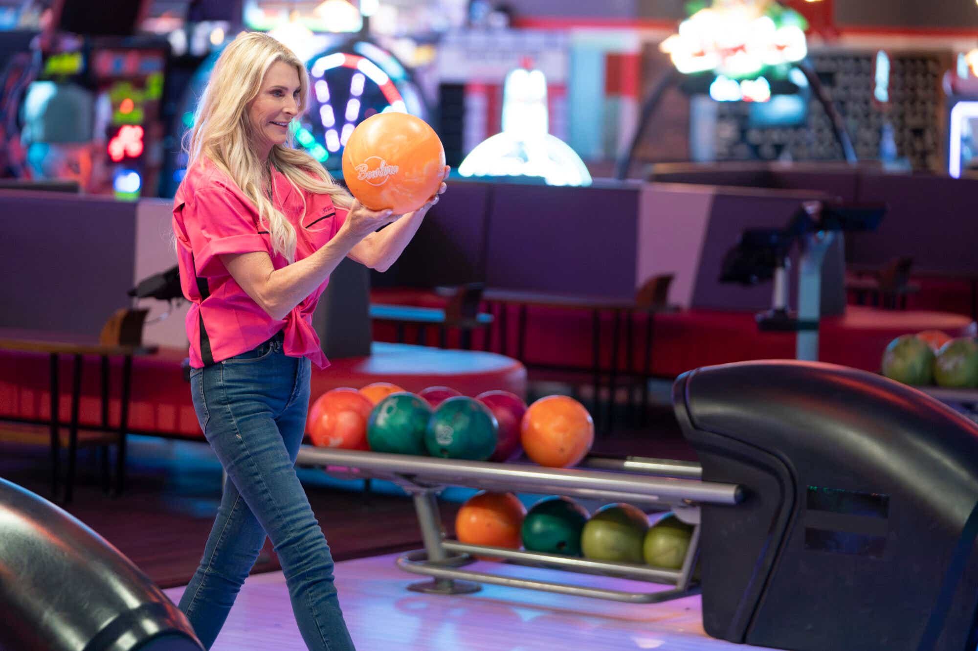 Joan Vassos wears a tied-up hot pink bowling shirt and jeans and gets ready to bowl a bright orange ball.