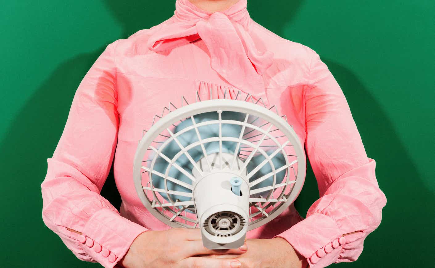 Woman in pink shirt with sweat stains holds fan in front of her