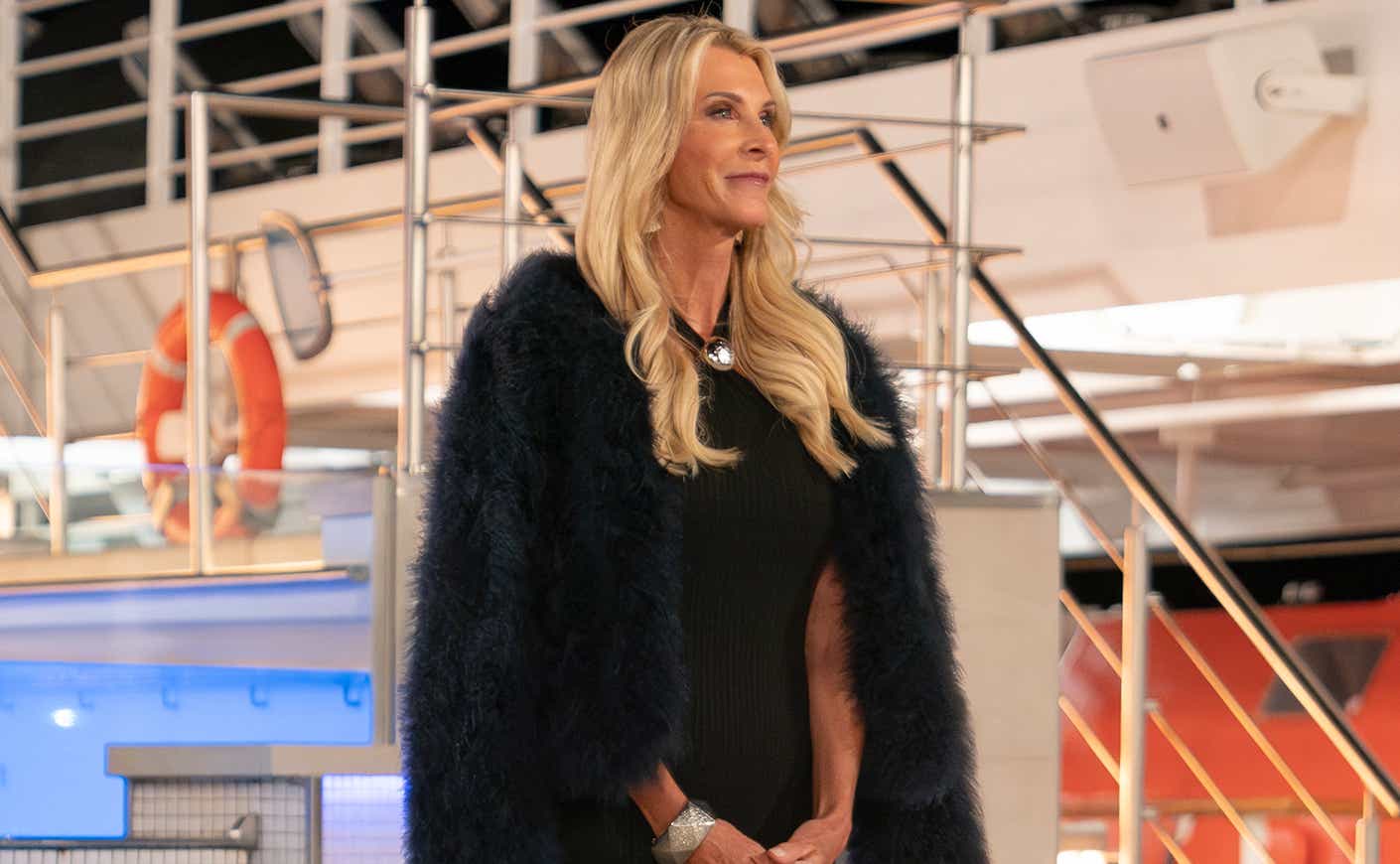 Joan Vassos stands on the deck of a ship in a black dress with a black furry jacket draped over her shoulders.