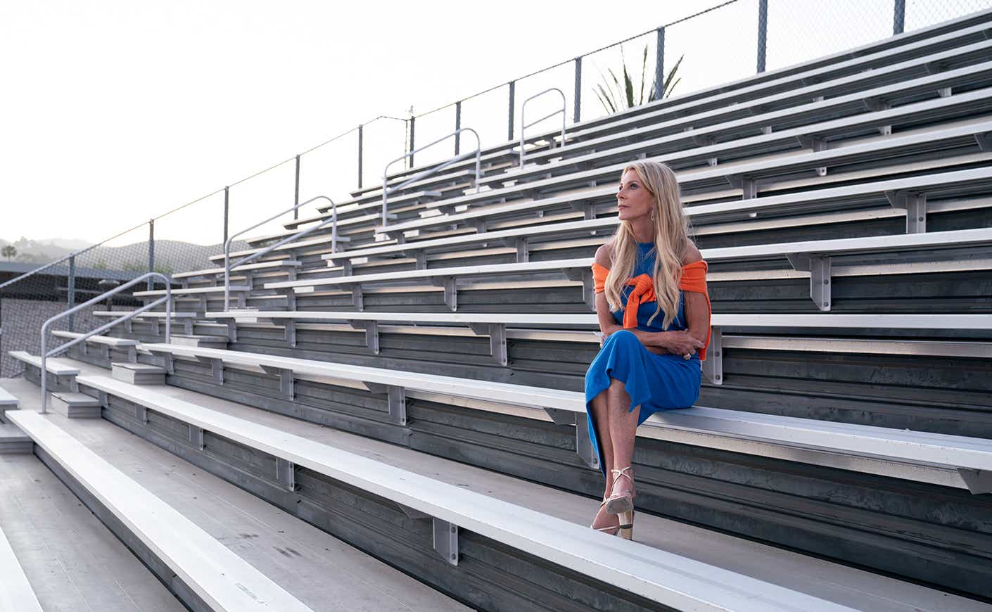 Joan Vassos wears a blue dress and orange sweater tied around her shoulder, sitting on bleachers, deep in thought.