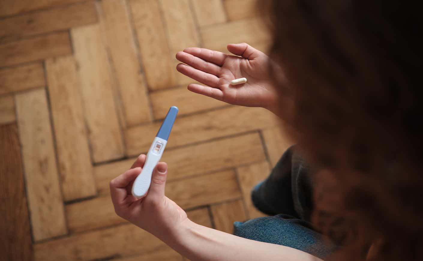 Woman holding pregnancy test and abortion pill while sitting on sofa.