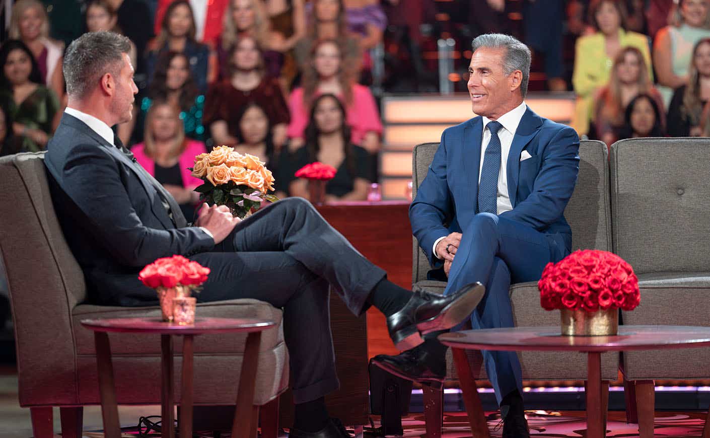 Guy at After the Final Rose, sitting on a gray couch wearing a navy blue suit. He's talking to Jesse Palmer, who's wearing a dark gray suit and sitting on an armchair perpendicular to Guy.