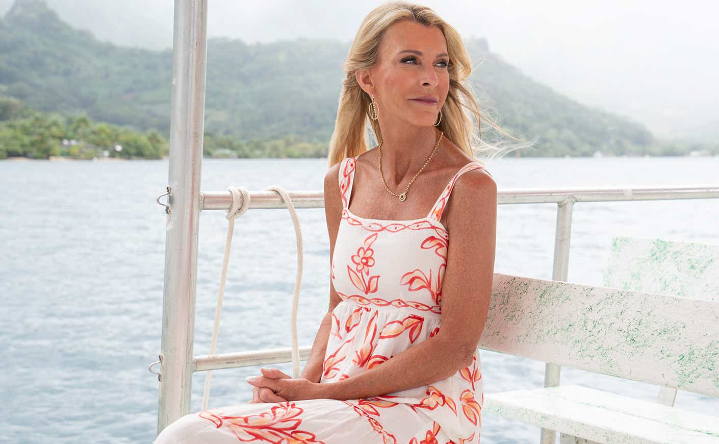 Joan Vassos sits on a boat, staring out at the ocean, wearing a white dress with a coral print