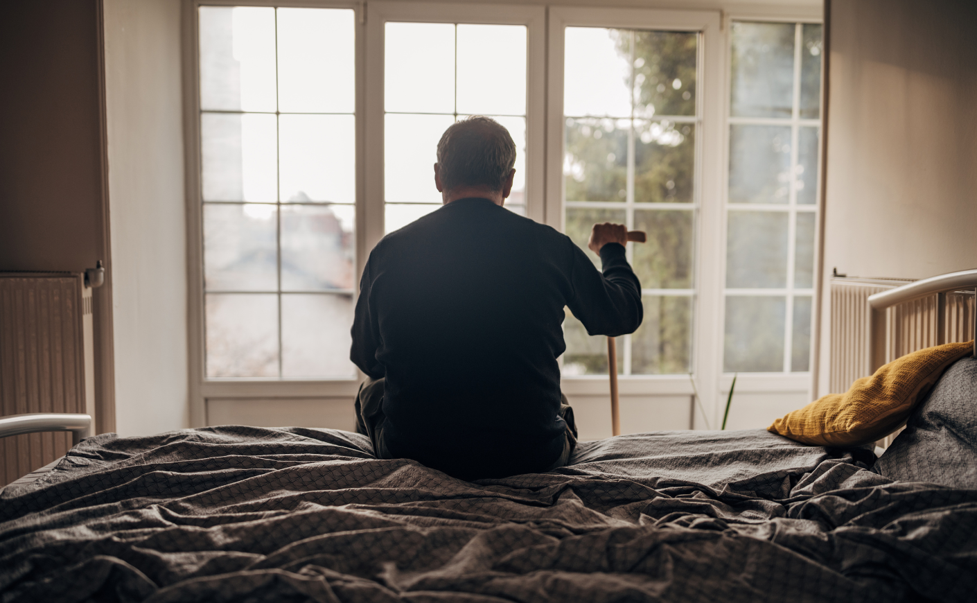 Elderly man sitting on a bed, holding a cane, looking out the window