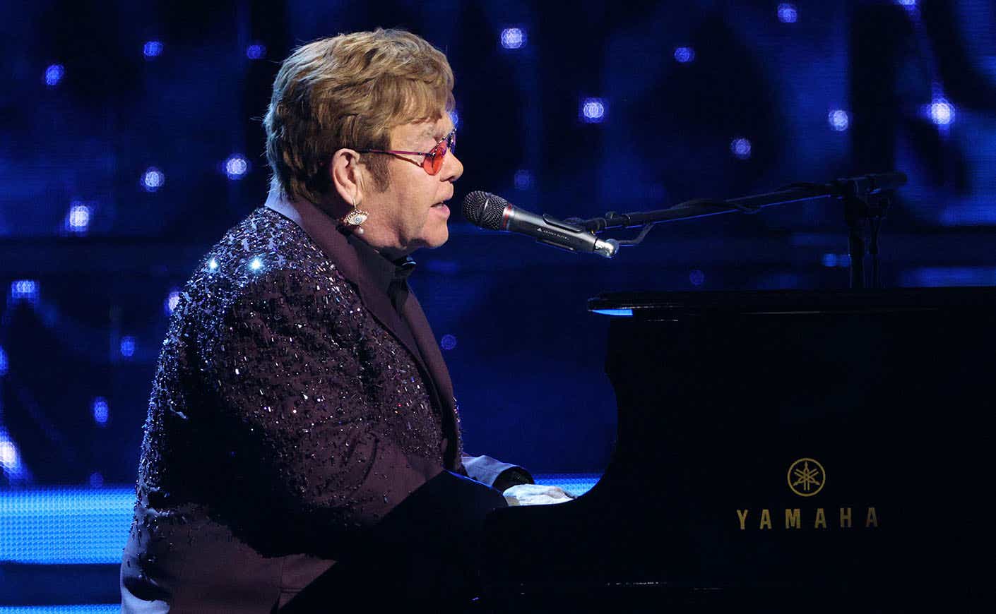 Elton John wearing a bejeweled purple blazer playing the piano