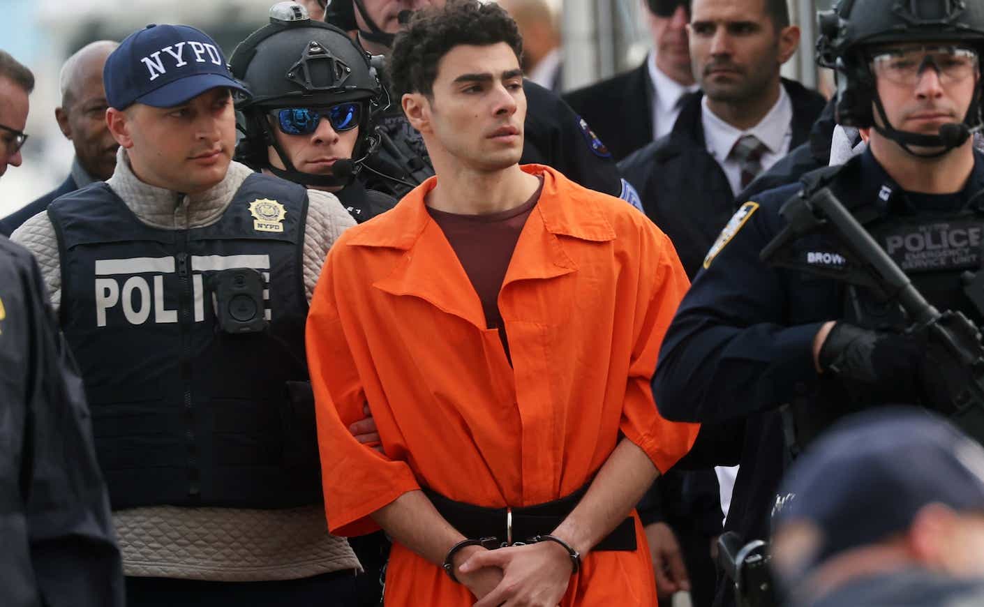 luigi mangione in an orange jumpsuit and handcuffs being escorted by armed guards