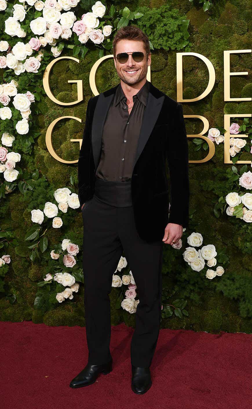 Glen Powell arrives at the Golden Globes in a black suit and a grayish-brown shirt, no tie, sunglasses.