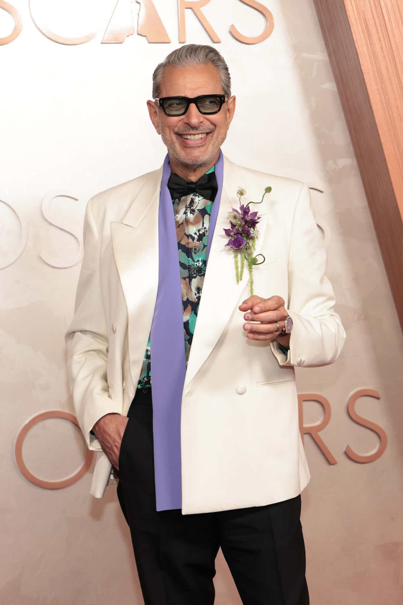 Jeff Goldblum wears a white blazer with a teal, brown and black floral patterned shirt underneath, a black bowlie, and a lavender scarf. He has a purple flower boutenniere.