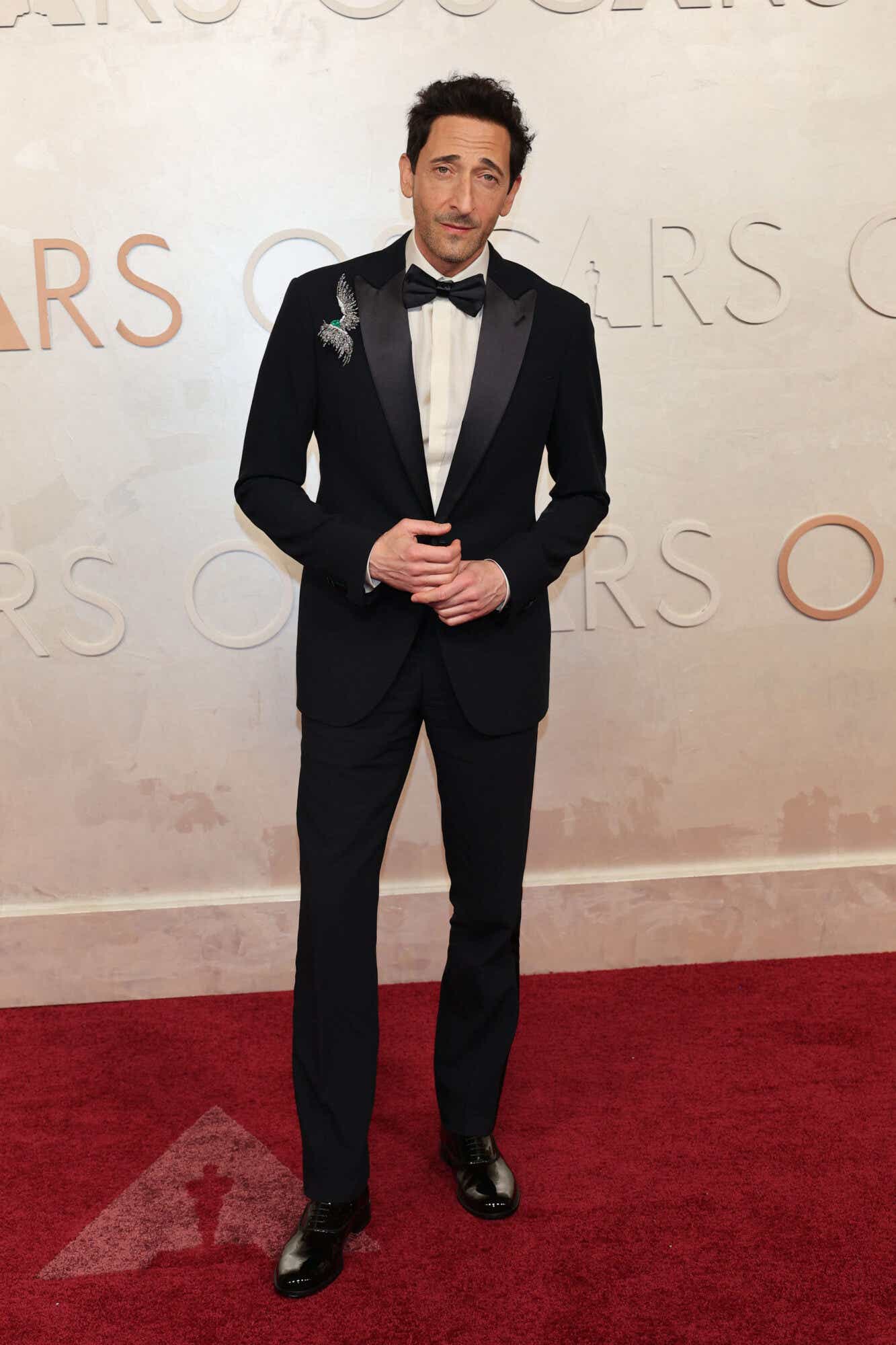 Adrien Brody wears a black tux with a pin depicting a bird in flight on his lapel.