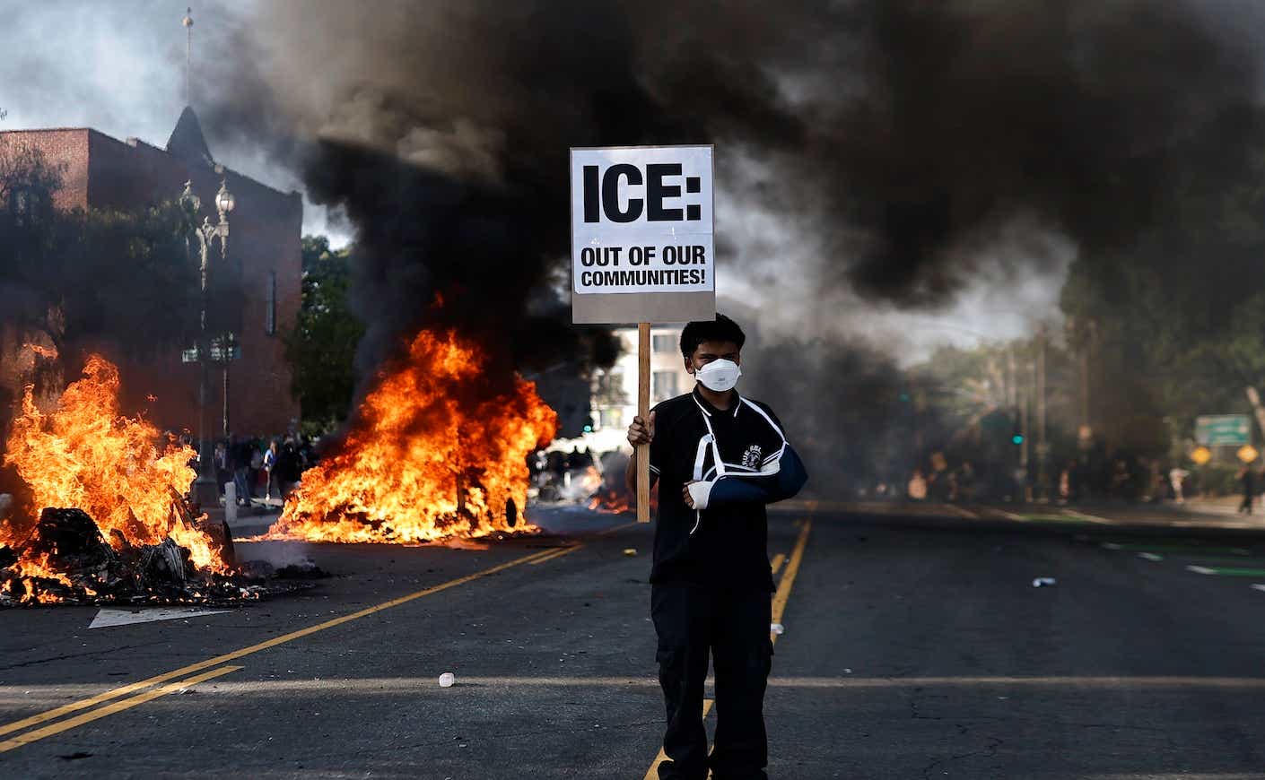a man in a mask holding protest sign in front of fires in LA