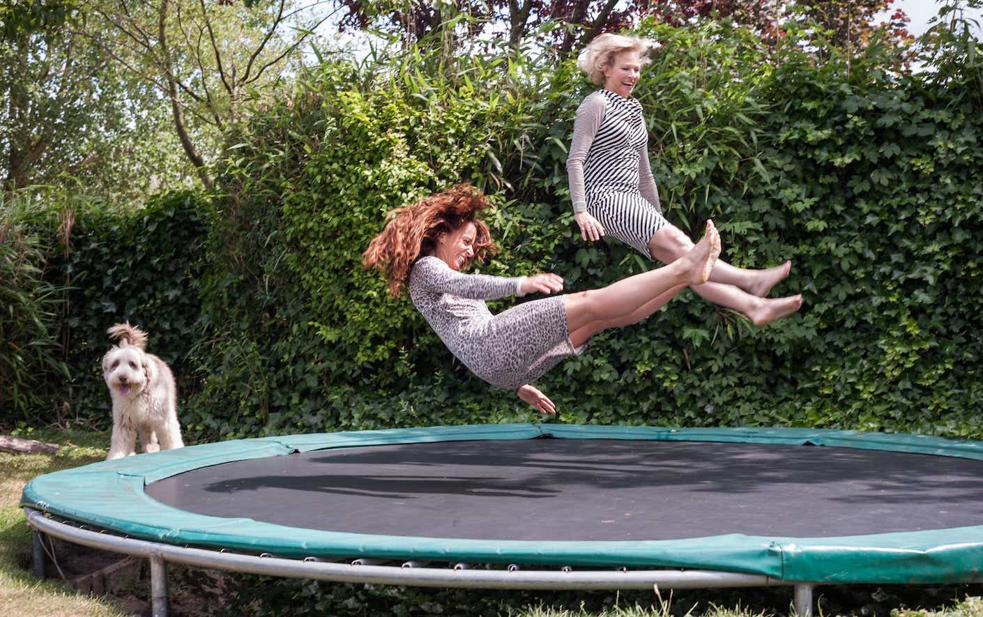 Two older women jumping on trampoline with a dog in the background
