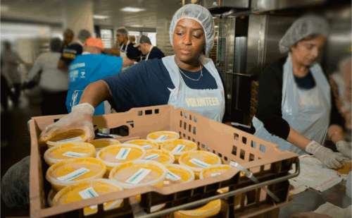 volunteer woman in an apron in a kitchen putting containers of soups in a crate.
