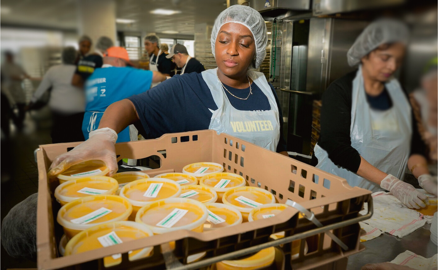 volunteer woman in an apron in a kitchen putting containers of soups in a crate.