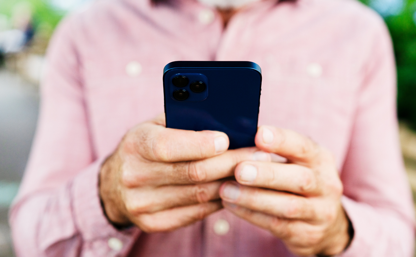 close-up of elderly man in a pink button-down typing on his black iPhone