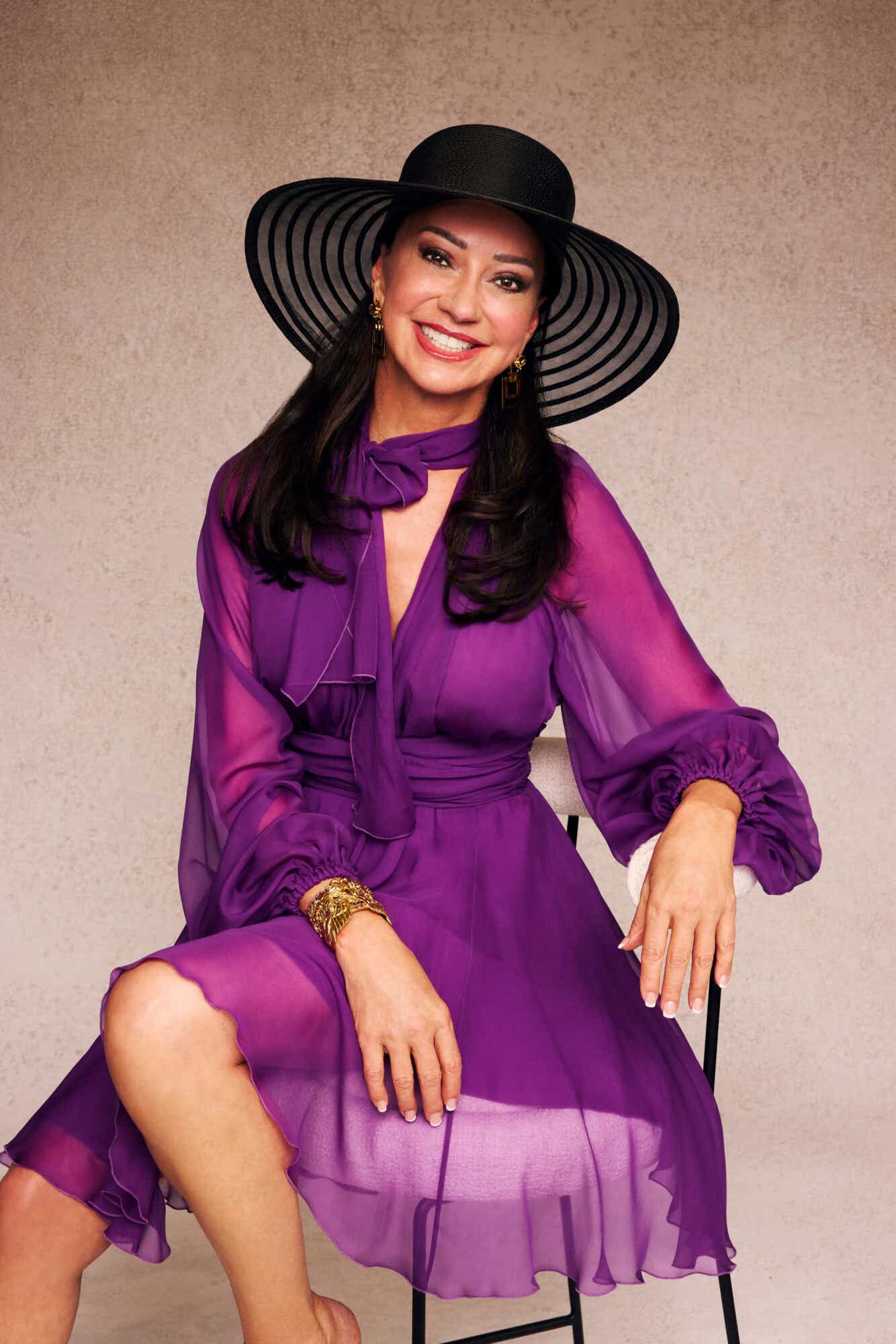 Tracy wears a flowy long-sleeve purple dress and a big black hat