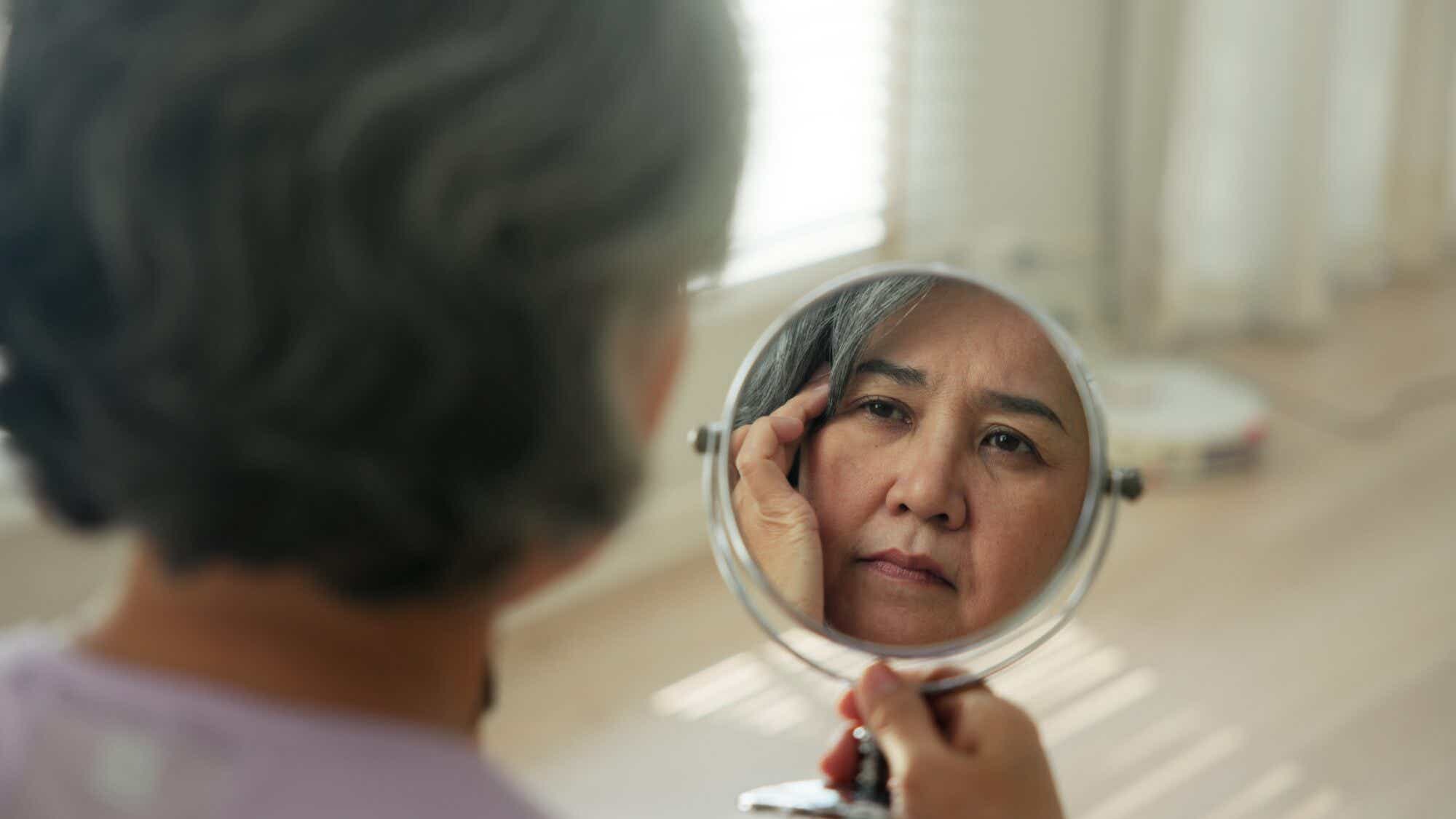 An Asian woman with short gray hair looks at her reflection in a hand mirror