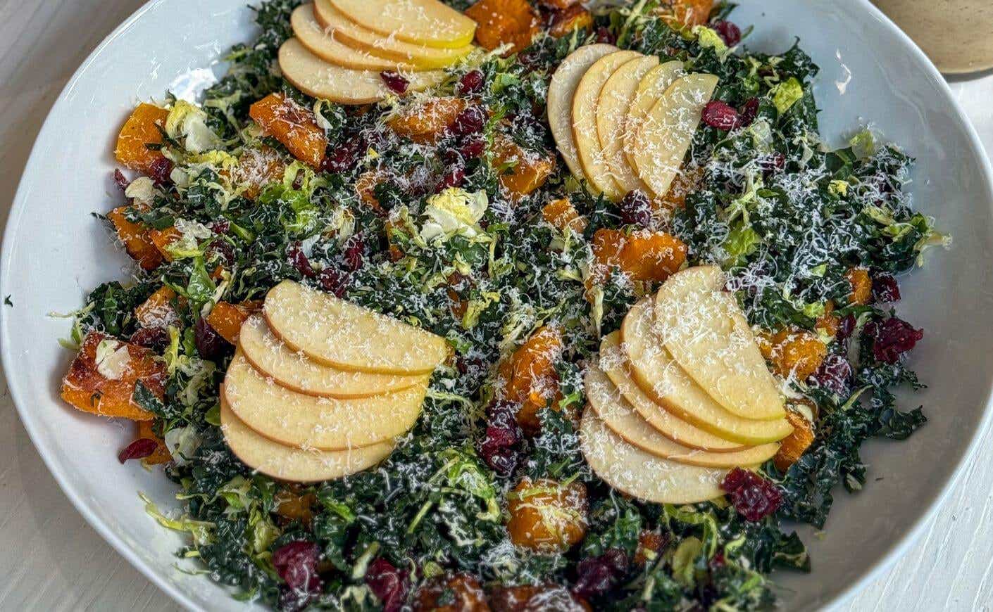 A bowl of kale, squash, and apple salad in walnut dressing and covered in parmesan cheese.