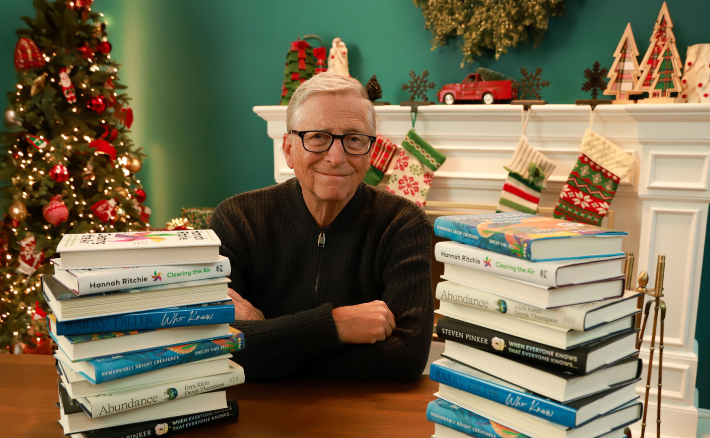 Bill Gates sitting between 2 stacks of books, there is a Christmas tree in the background