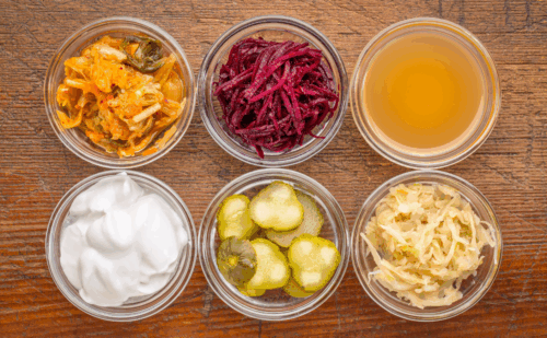 a set of fermented food great for gut health - top view of glass bowls against grunge wood: cucumber pickles, coconut milk yogurt, kimchi, sauerkraut, red beets, apple cider vinegar
