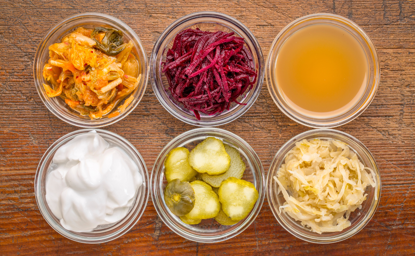 a set of fermented food great for gut health - top view of glass bowls against grunge wood: cucumber pickles, coconut milk yogurt, kimchi, sauerkraut, red beets, apple cider vinegar