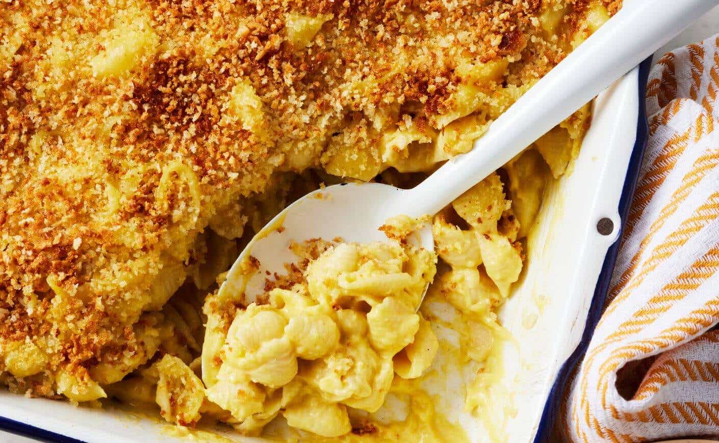 A serving spoon scooping from a baking pan of butternut squash mac and cheese with a crunchy topping.
